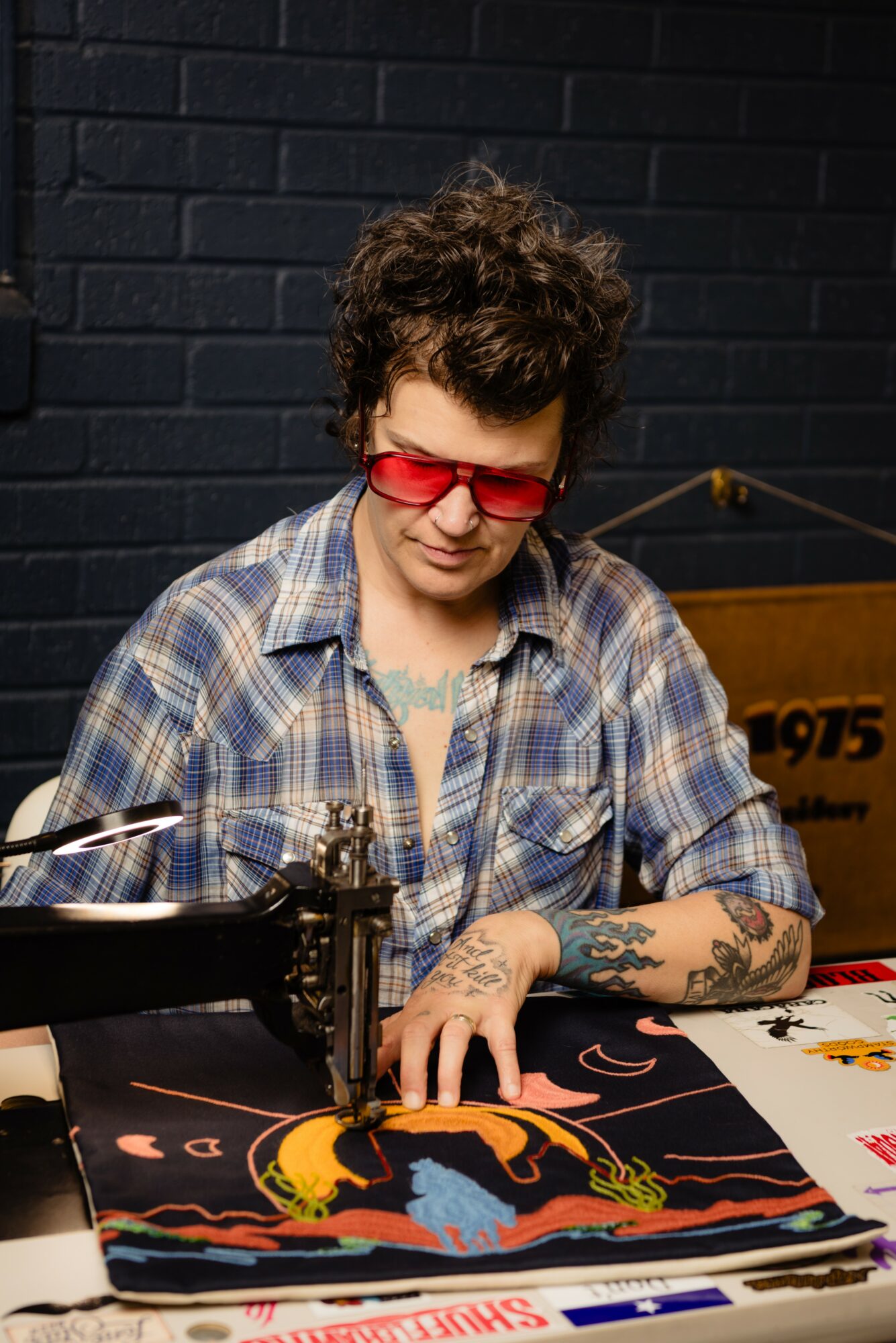 Person with curly hair and tattoos wearing red glasses and plaid shirt working on fabric with a sewing machine, black brick wall background.