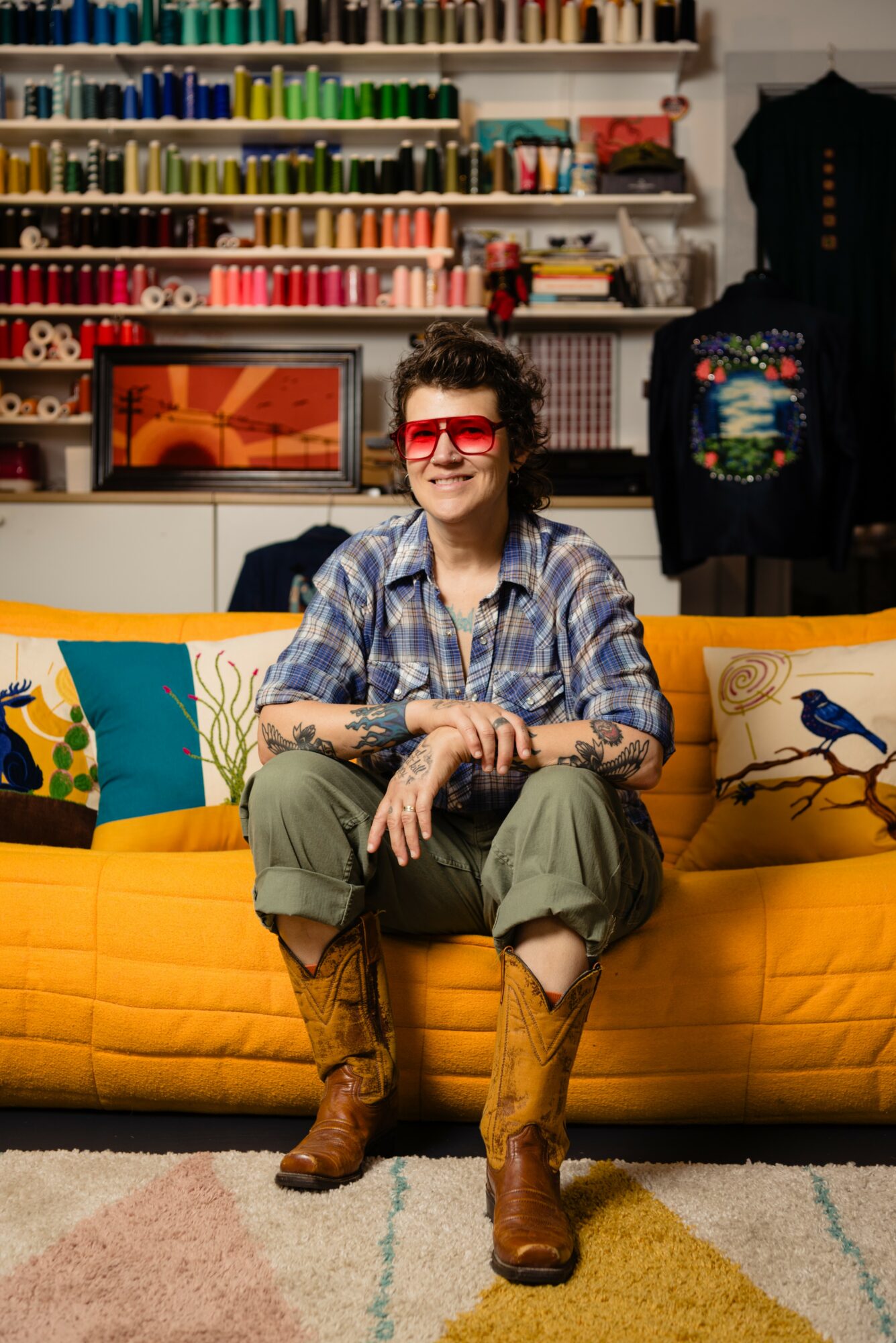 Person with curly hair and red glasses sitting on a yellow couch in a colorful room with books and clothing displays