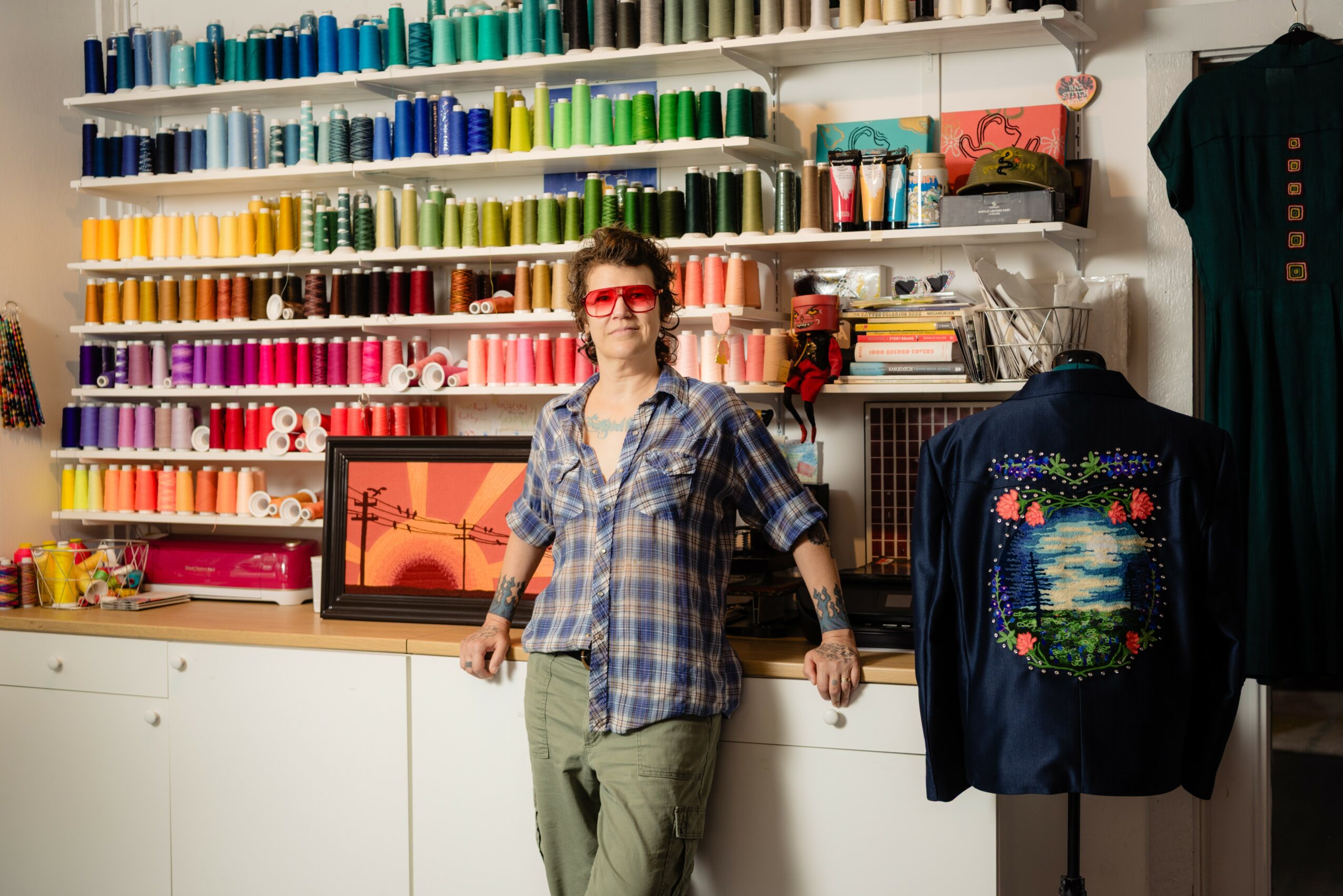 Person standing in front of shelves filled with colorful spools of thread, wearing glasses and a plaid shirt, smiling.