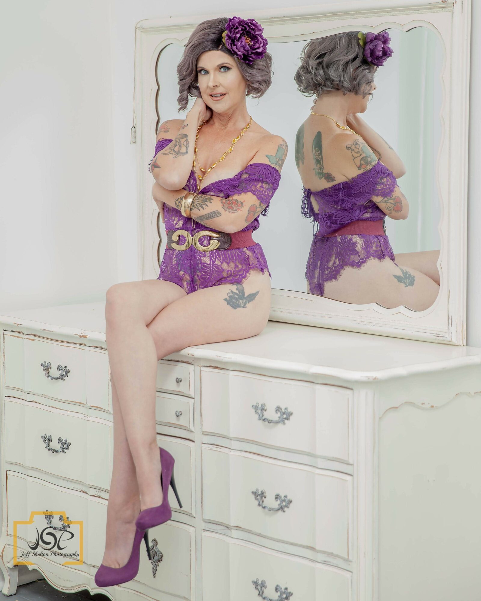 Woman with gray hair and purple flowers in hair, wearing purple lace lingerie and high heels, sitting on a white dresser, reflected in mirror.