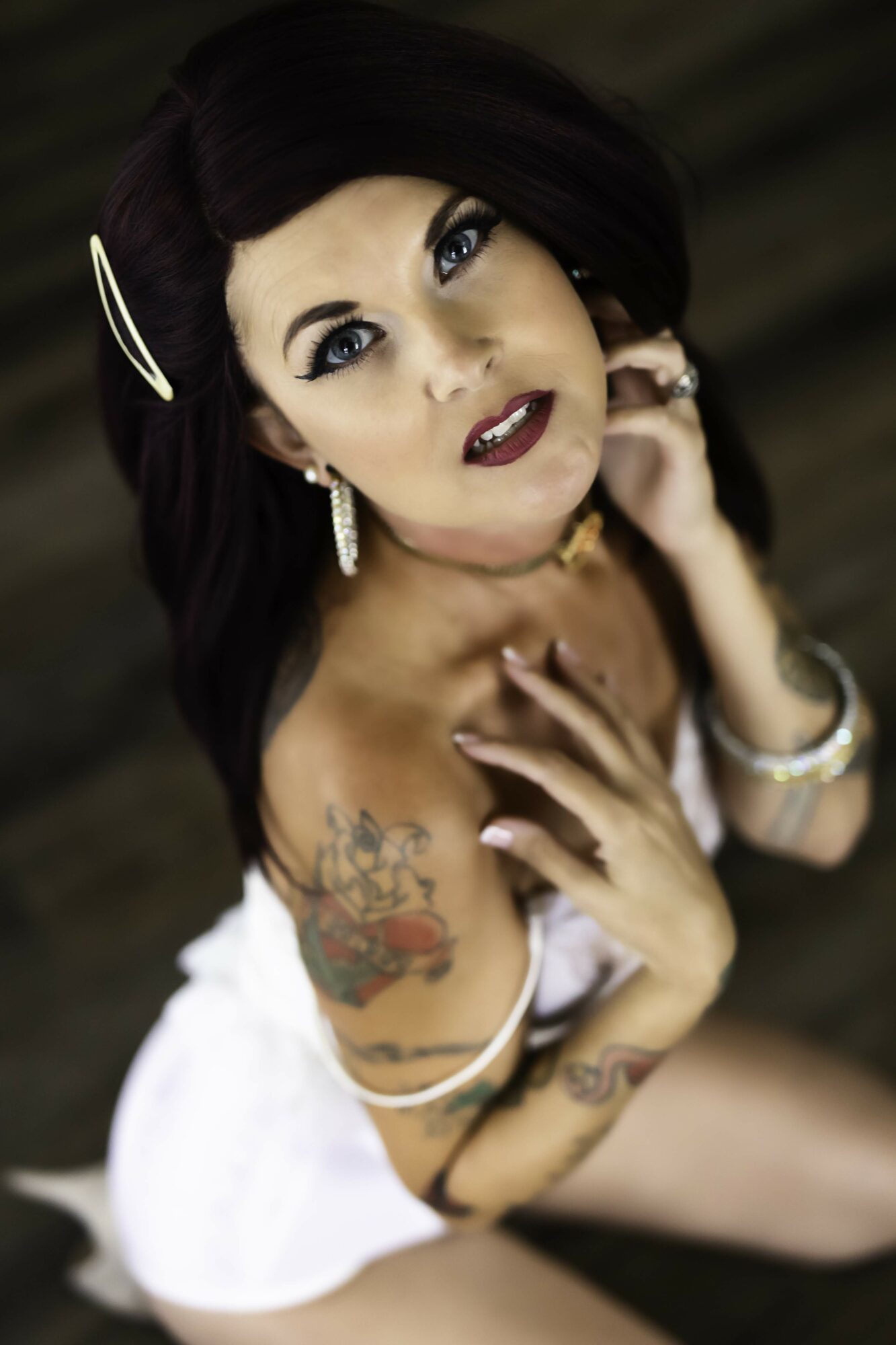 Woman with dark hair, makeup, tattoos, jewelry, and a white outfit, looking up at the camera with a blurred background.