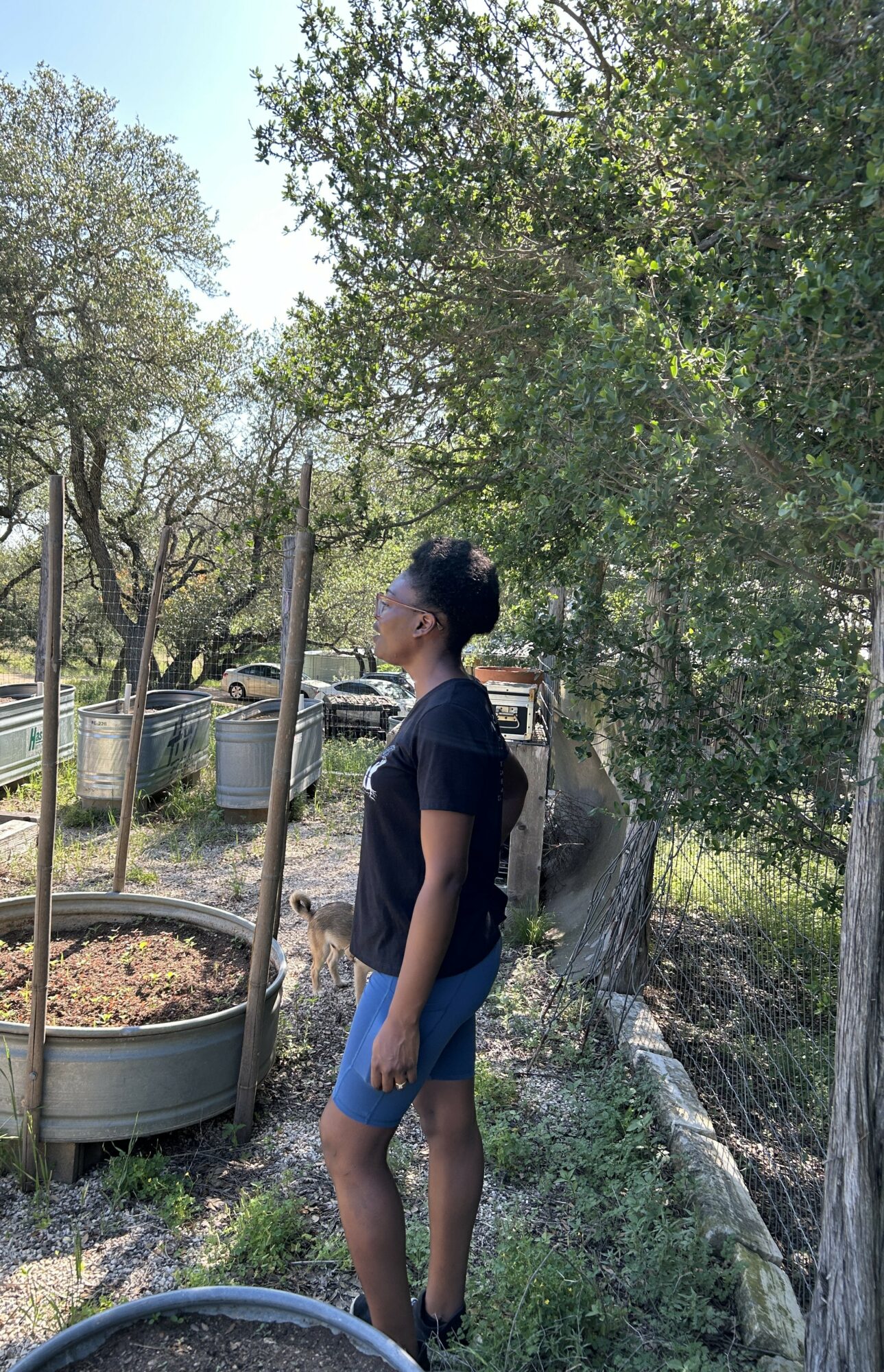 Young woman standing outdoors near trees and a garden bed, looking to the side, wearing a black shirt and blue shorts.