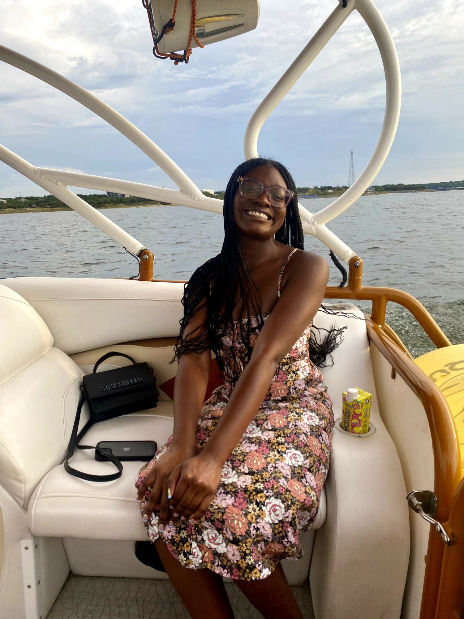 Smiling woman with long hair wearing a floral dress sitting on boat seat near water, with boat structure and sky in background.