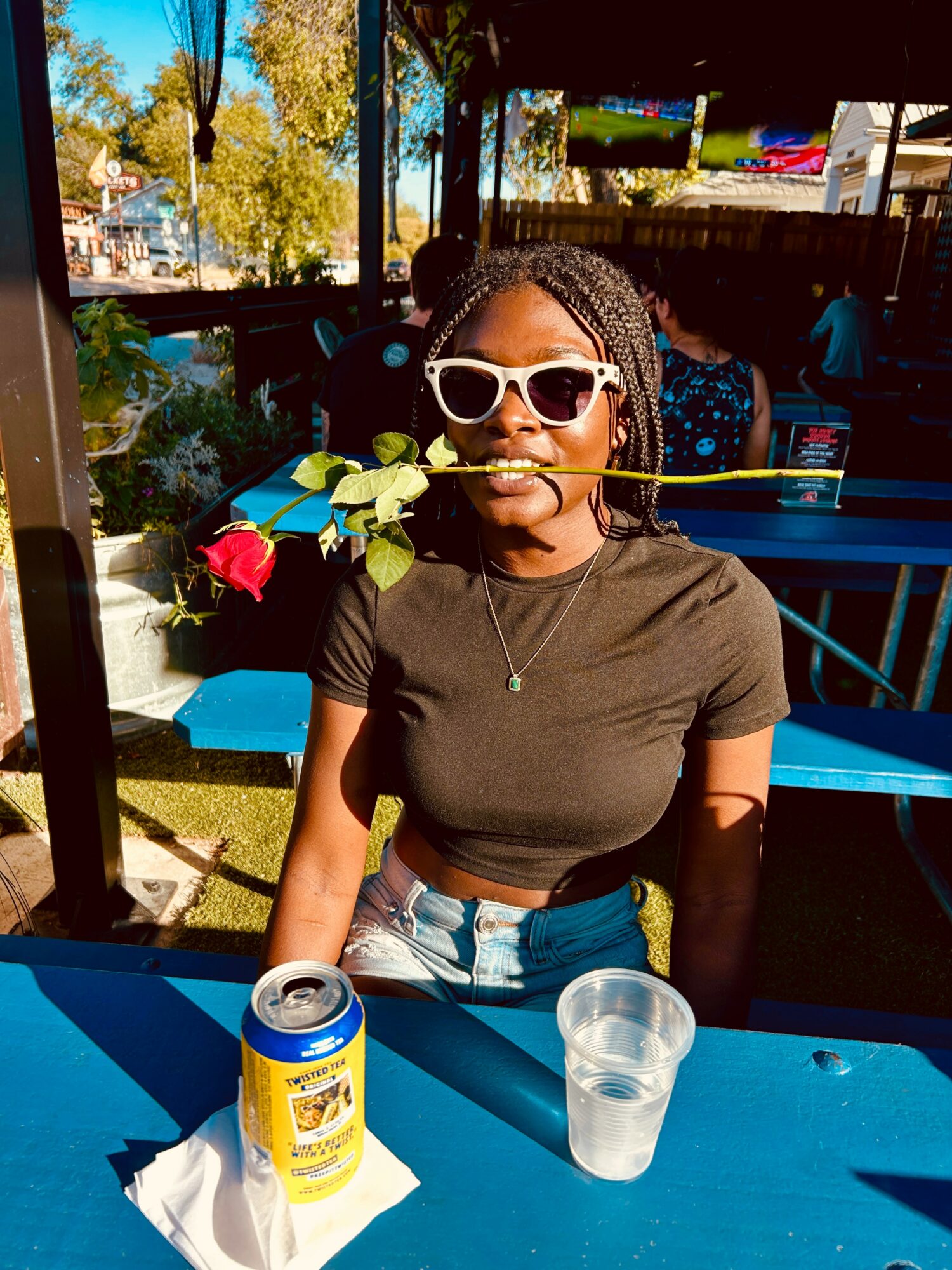 Young woman with sunglasses holding a rose in her mouth, sitting at a blue table outdoors, with a can and glass in front.