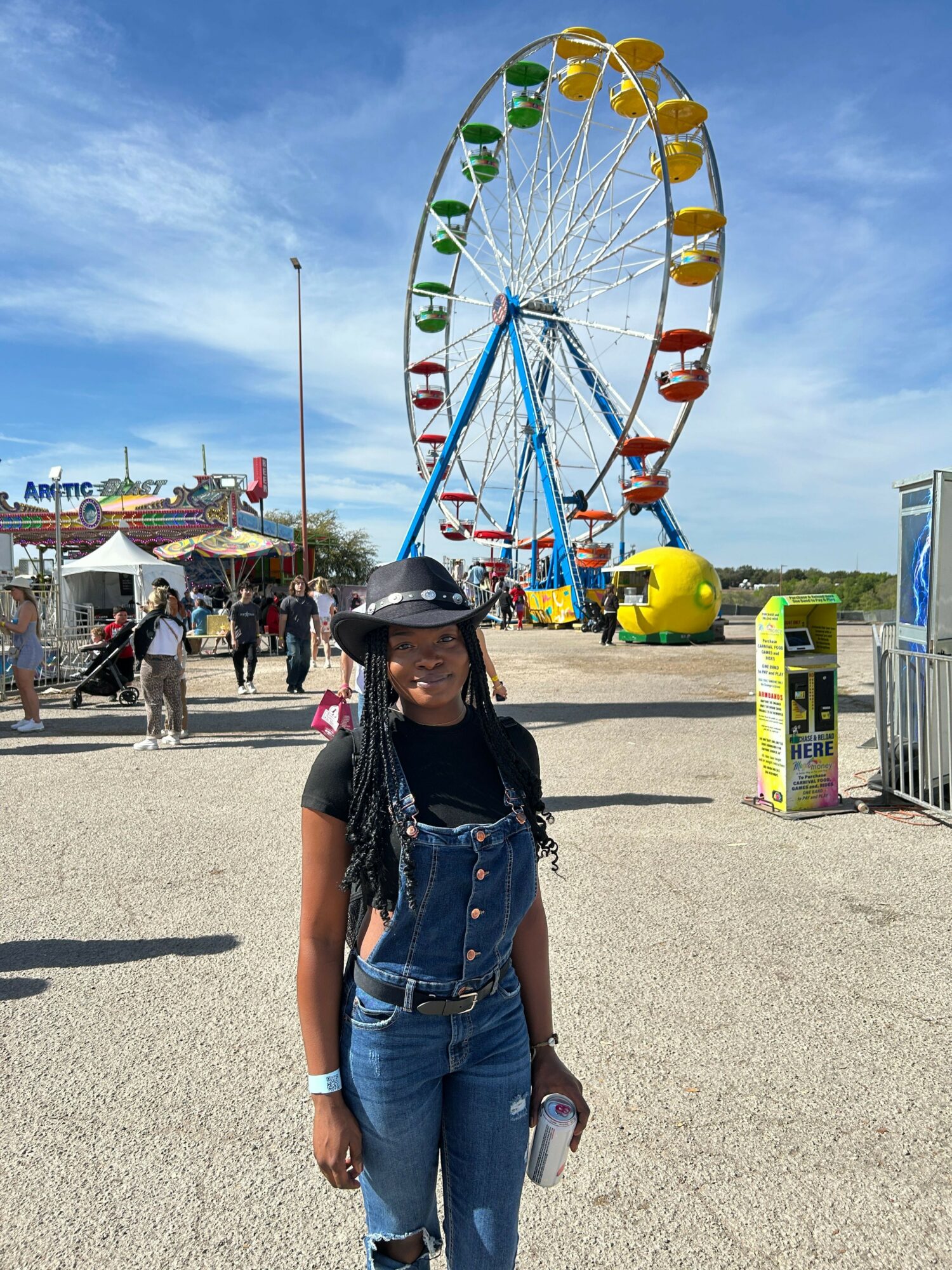 Person standing near a colorful Ferris wheel at an outdoor fair, with blue sky and other rides in background.