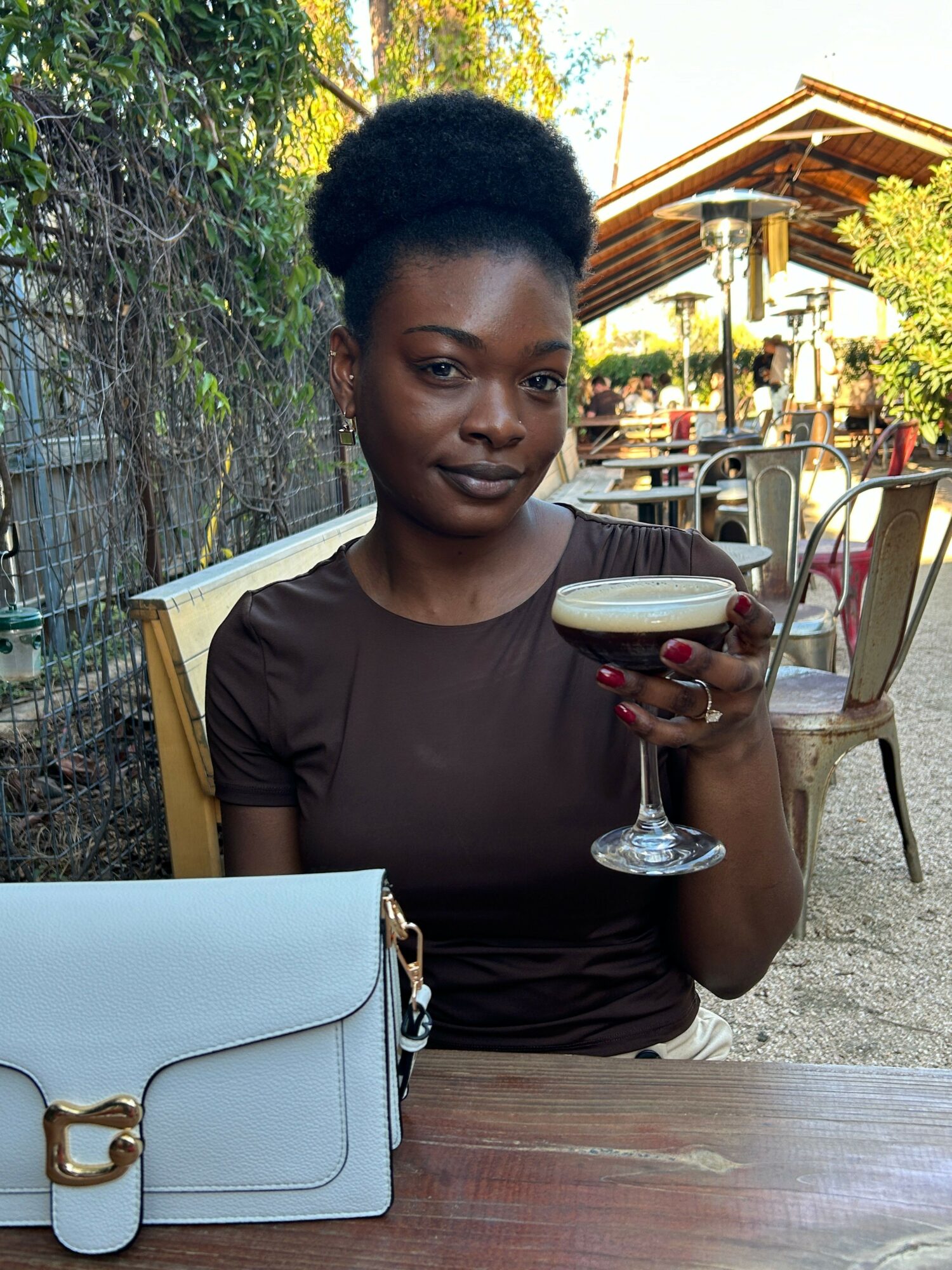 Woman sitting at a table holding a glass of wine outdoors, with a purse on the table, smiling at the camera.