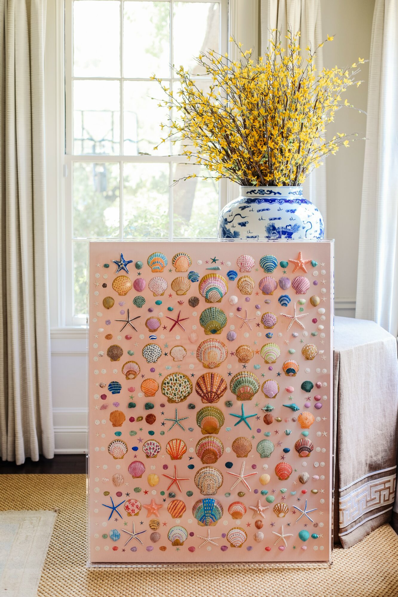 Colorful decorated board with seashells, starfish, and other beach-themed items, placed in front of a window with a vase of yellow flowers on top.