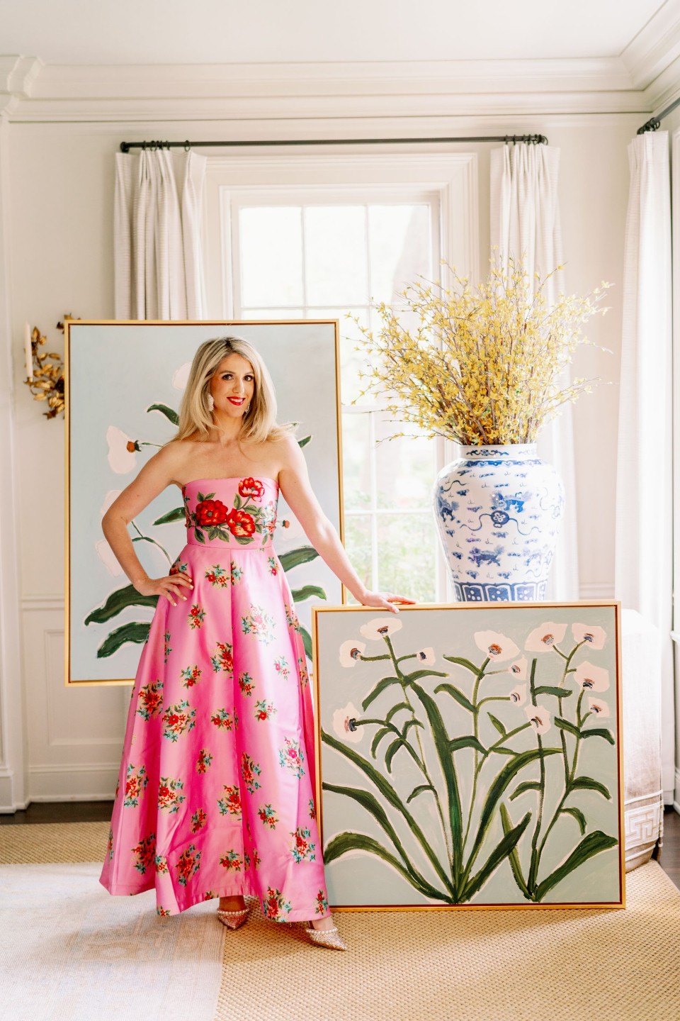 Woman in pink floral dress standing indoors with large framed artwork and a blue and white vase with yellow flowers behind her.