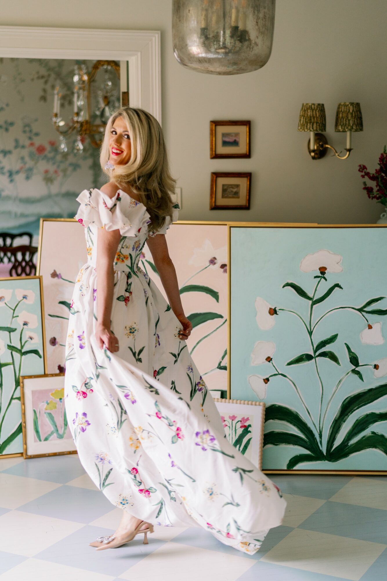 Woman in a floral dress standing indoors with framed artwork and decorative wall lights, smiling and looking to the side.