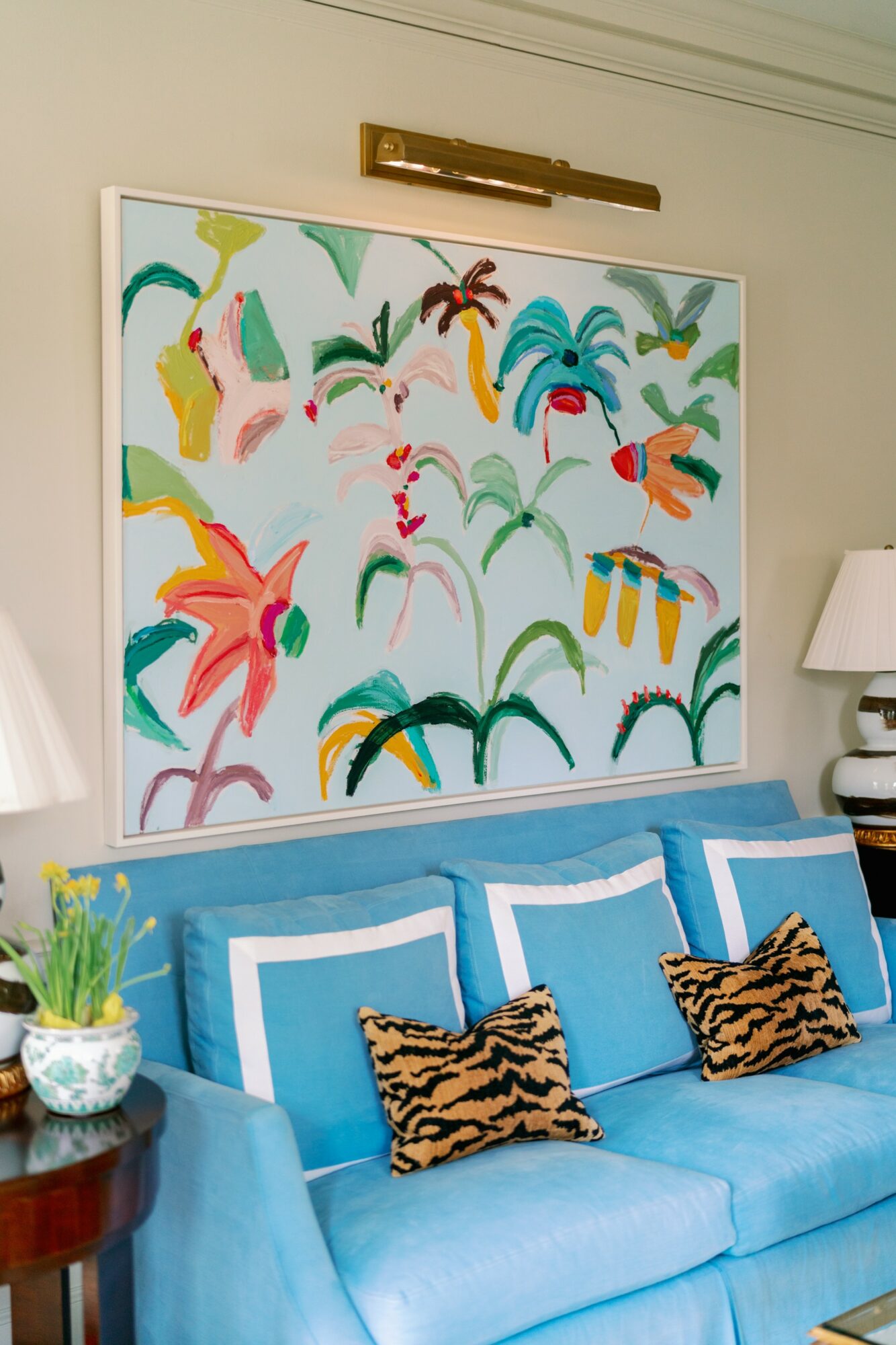 Colorful abstract painting hangs above a blue sofa with three pillows, a side table with a plant, and a lamp.