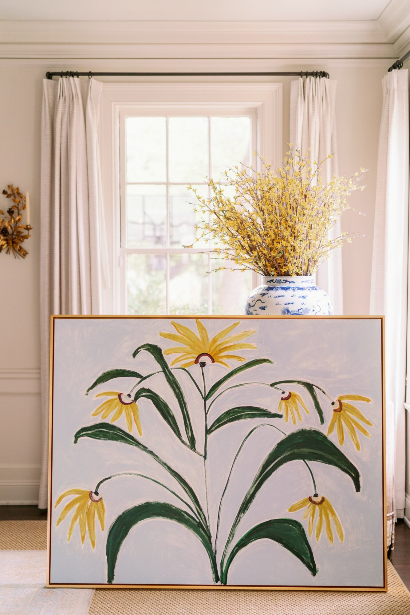 A large framed painting of yellow flowers with green leaves leans against a wall in front of a window with white curtains, with a vase of yellow flowers on the windowsill.
