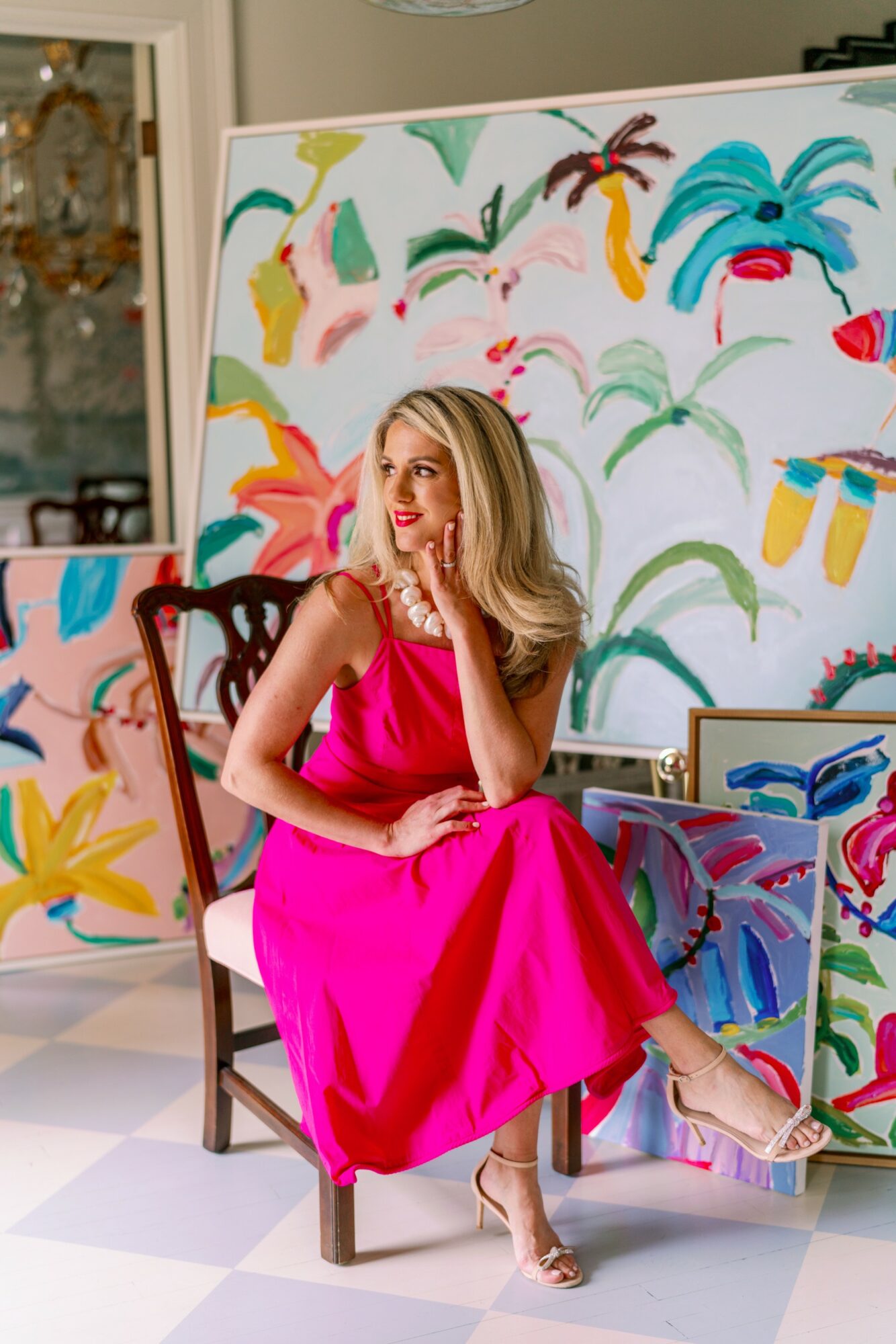 Woman in a pink dress sitting on a chair in front of colorful abstract paintings, smiling and looking to the side.