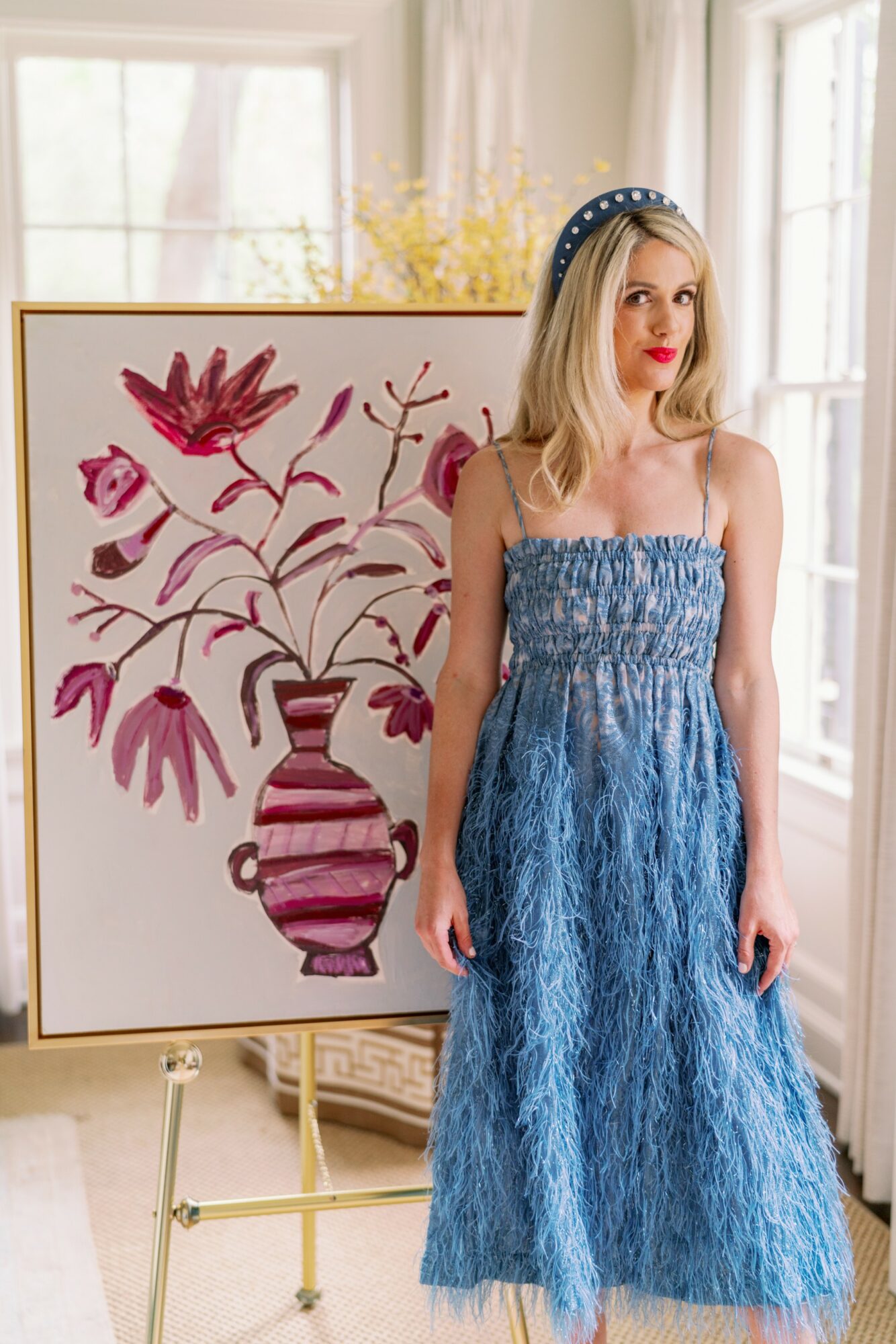 Young woman with blonde hair wearing a blue dress and headband, standing next to a large floral painting in a bright room.