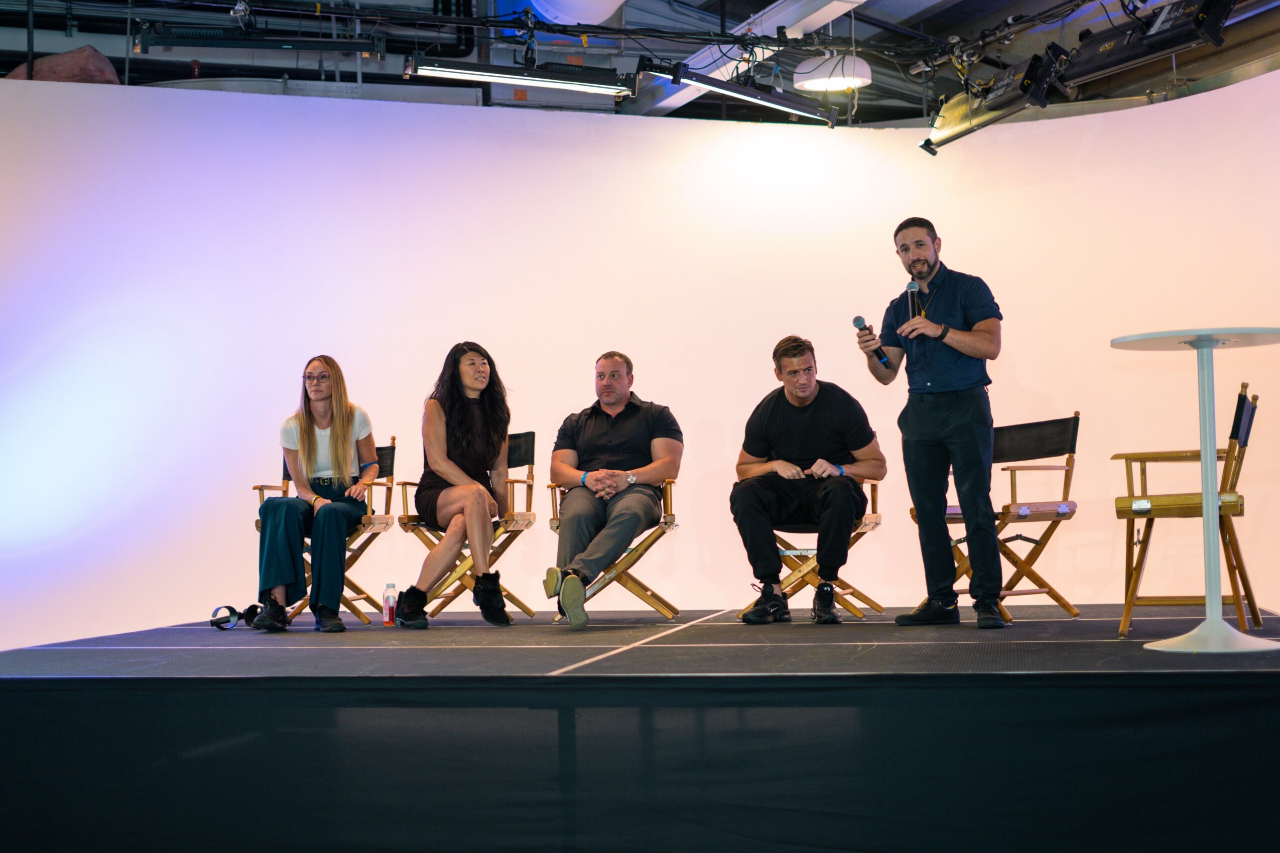 Five people on stage, four seated and one standing, with a plain background and minimal props.