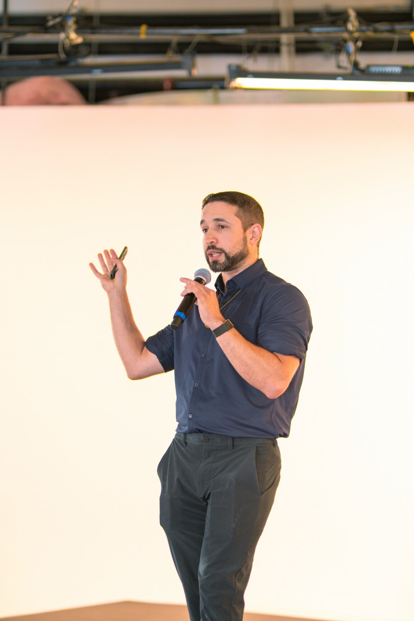 Man speaking into a microphone, gesturing with his left hand, wearing a dark shirt and pants, standing against a plain background.
