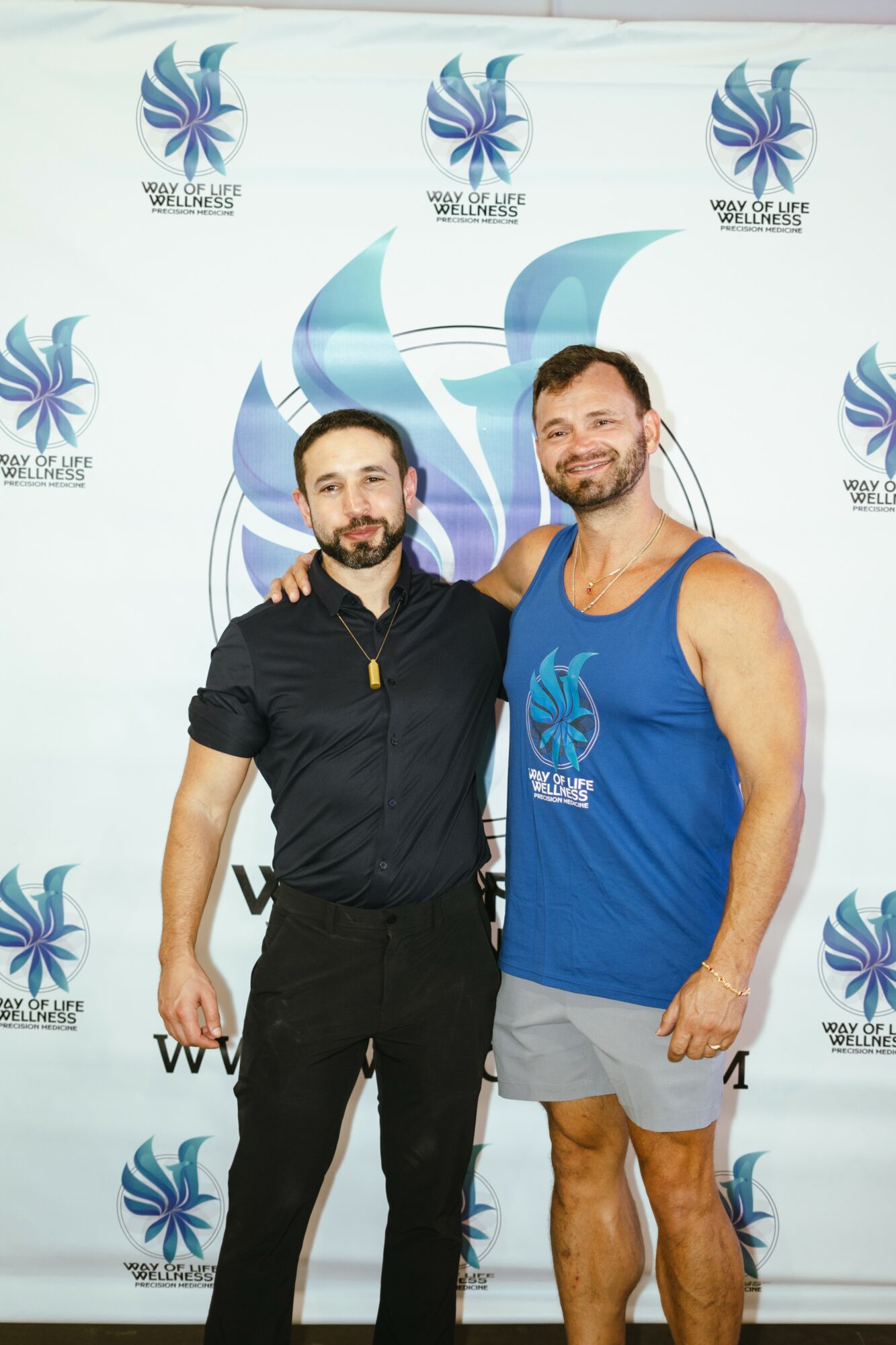 Two men standing together in front of a backdrop with a logo, one in a black shirt and the other in a blue tank top and shorts.