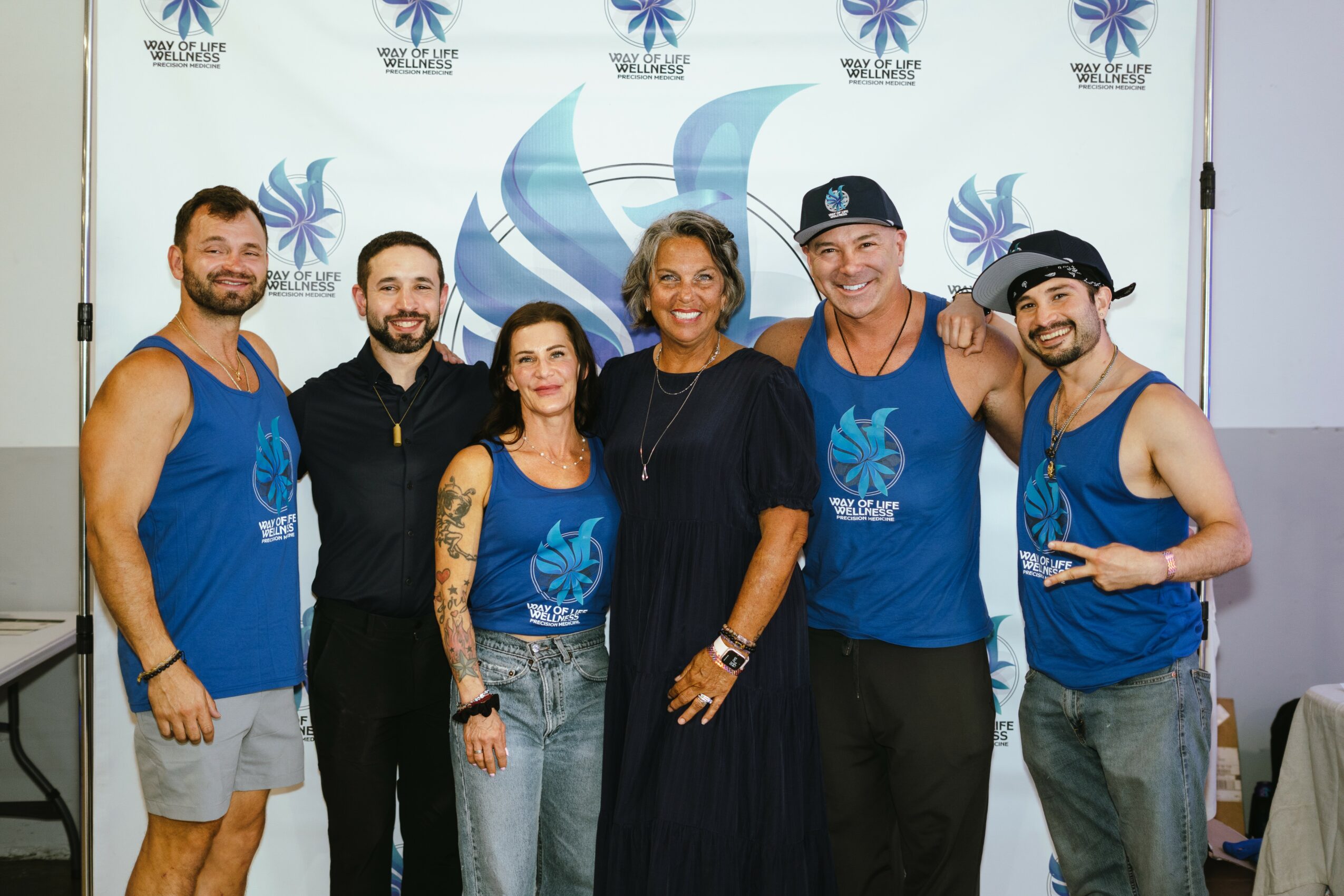 Group of six people smiling, standing in front of a backdrop with a logo, some wearing blue tank tops, one in black dress.