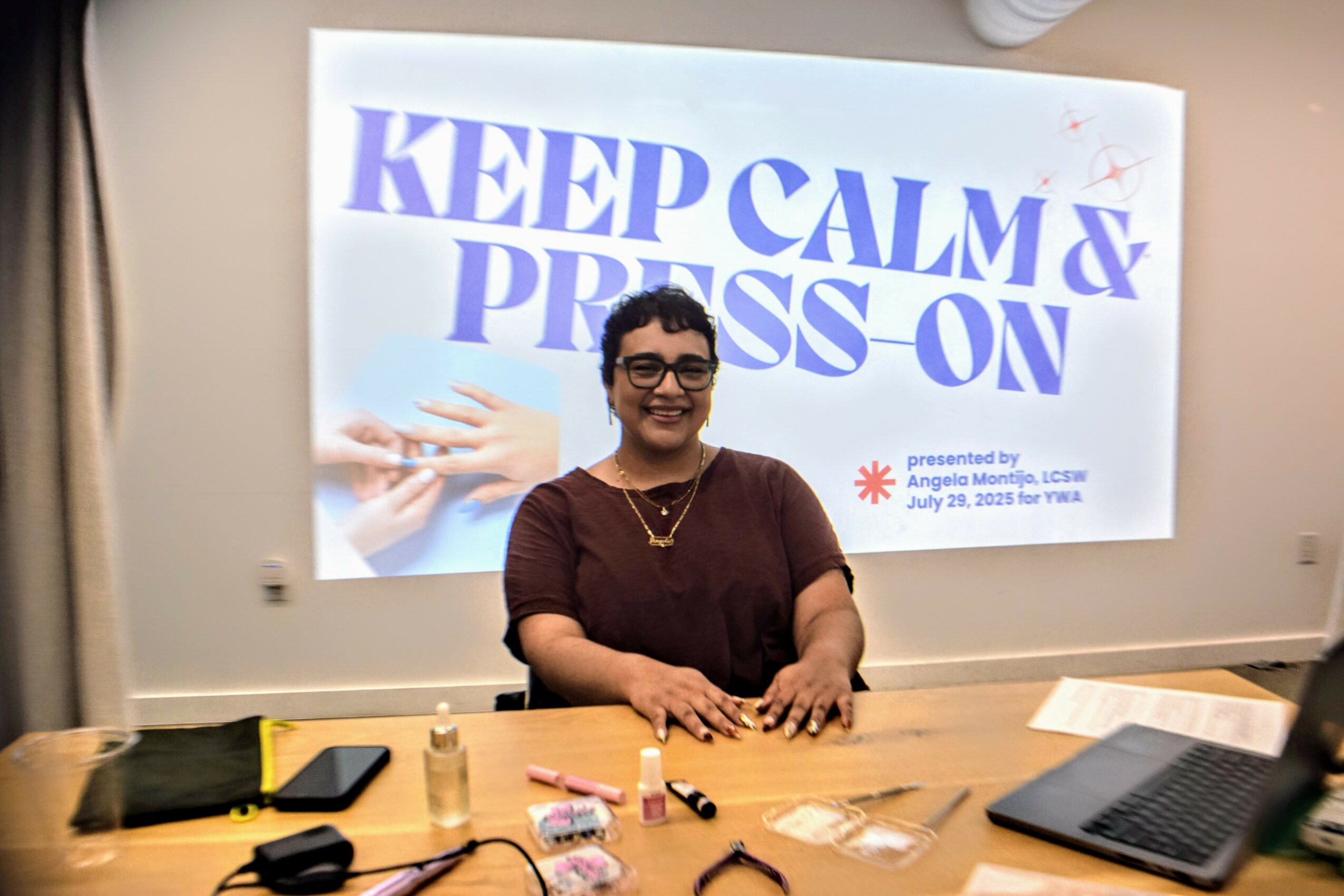 Woman sitting at a desk in front of a presentation screen, smiling, with a laptop and various items on the desk.