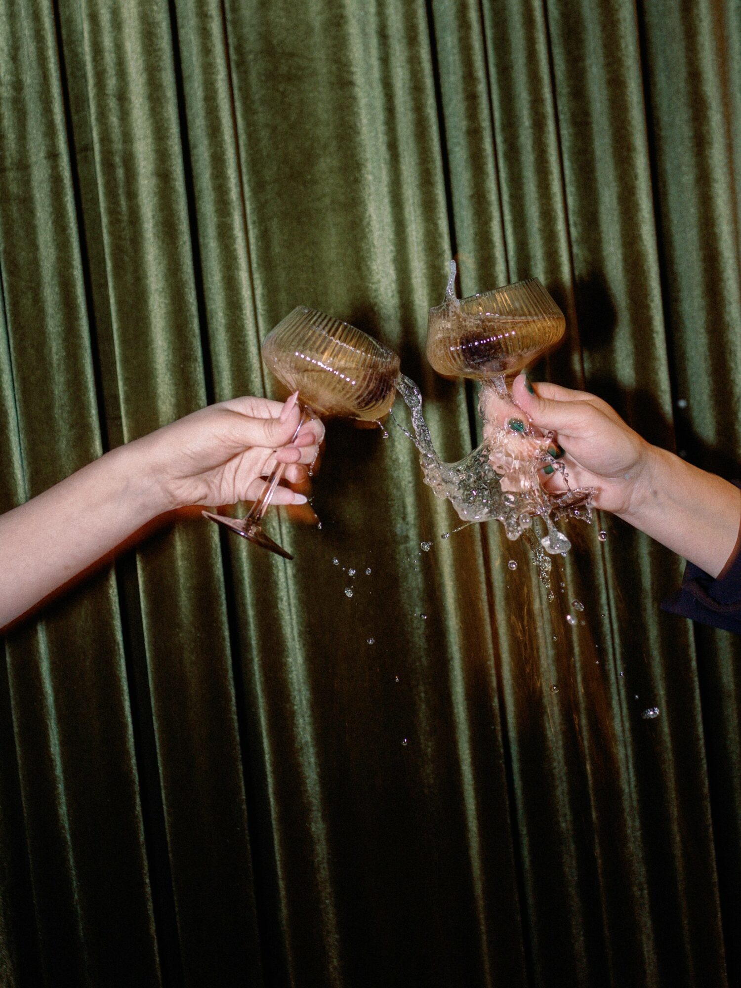 Two hands holding glasses with liquid, splashing against a green curtain background.