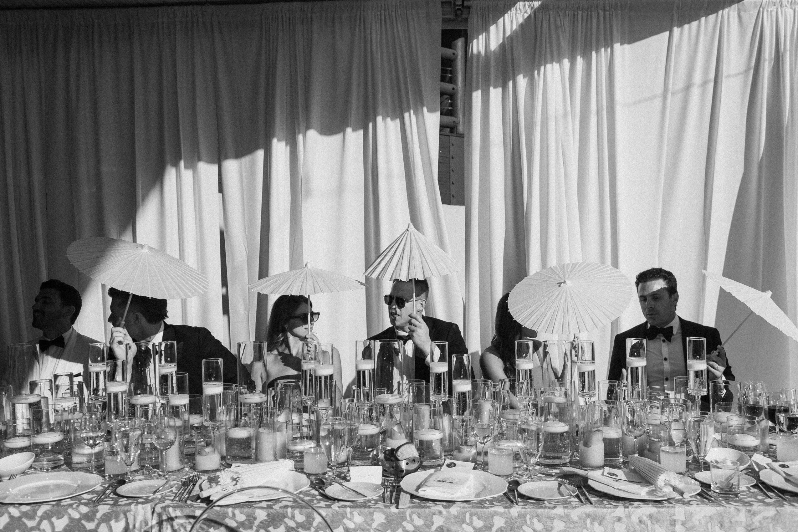People sitting at a table with umbrellas and drinks, with curtains in the background.