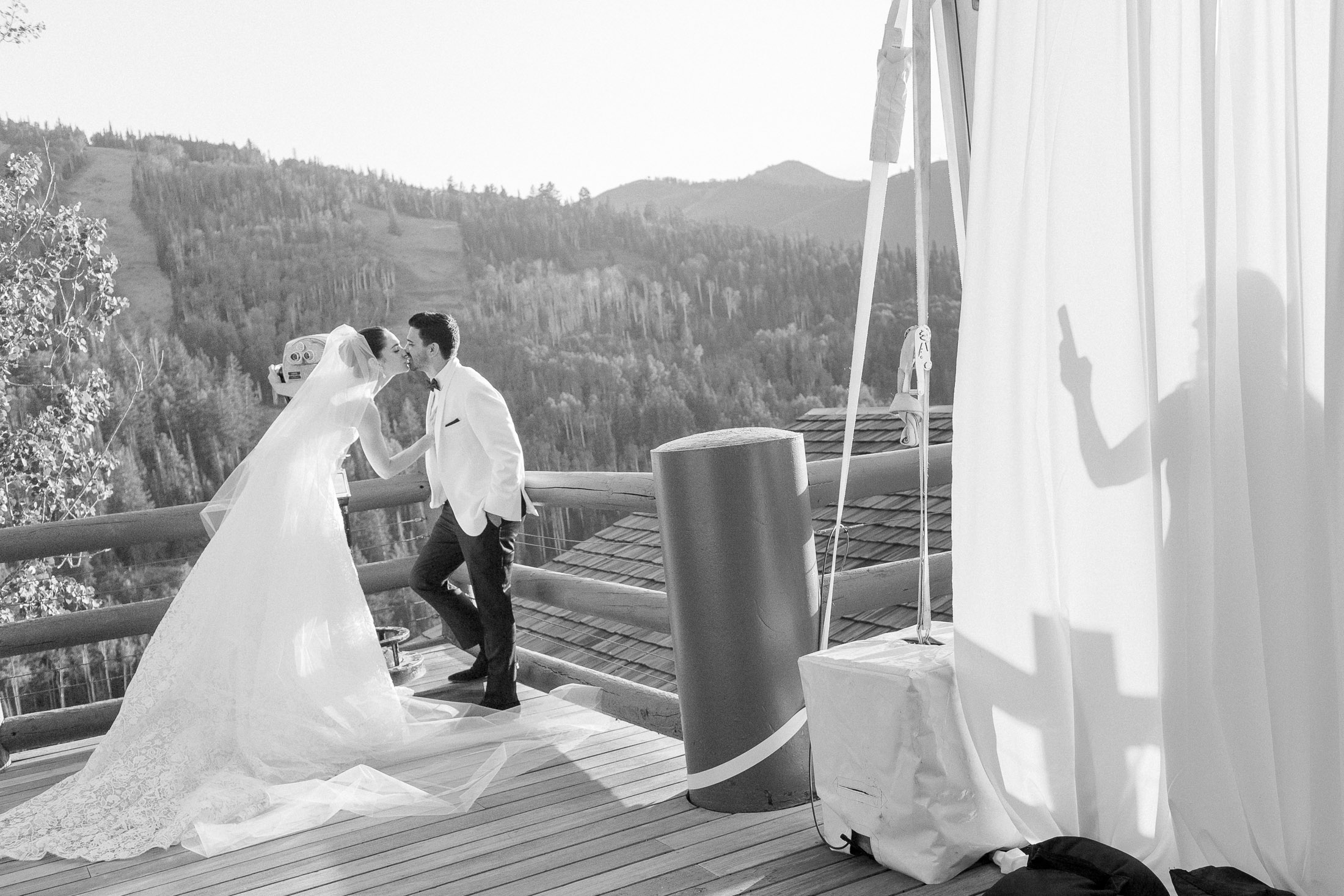 Bride and groom holding hands on a balcony with scenic mountains in the background, shadow of a person on a curtain, wedding scene.