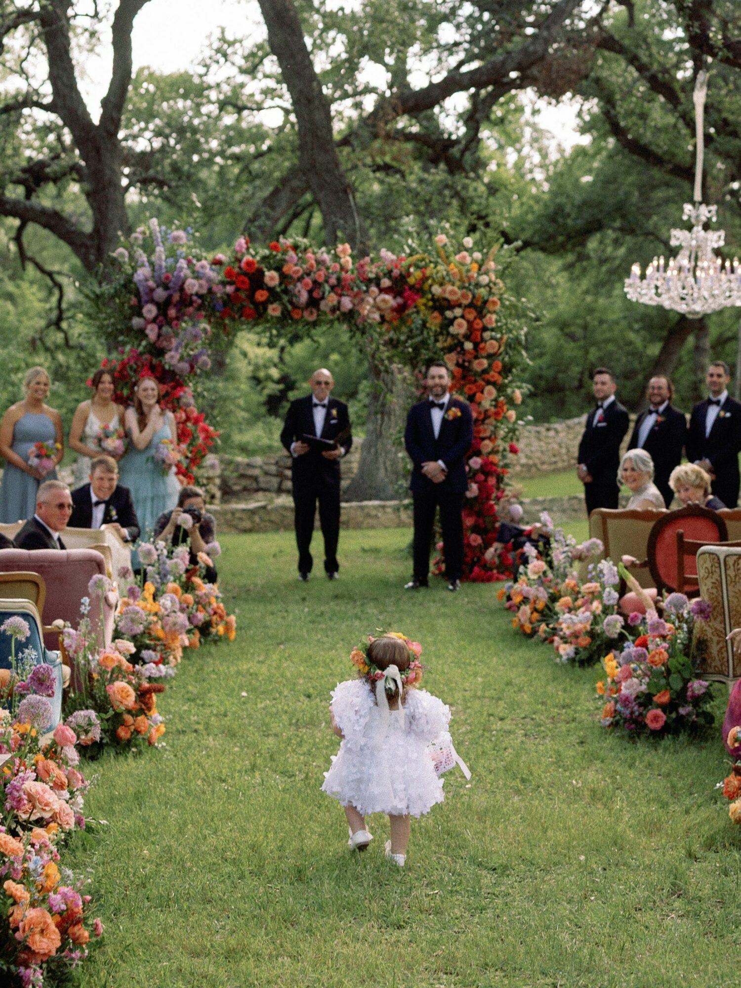 Child in white dress walking toward a wedding ceremony with floral arch, guests, and trees in background.