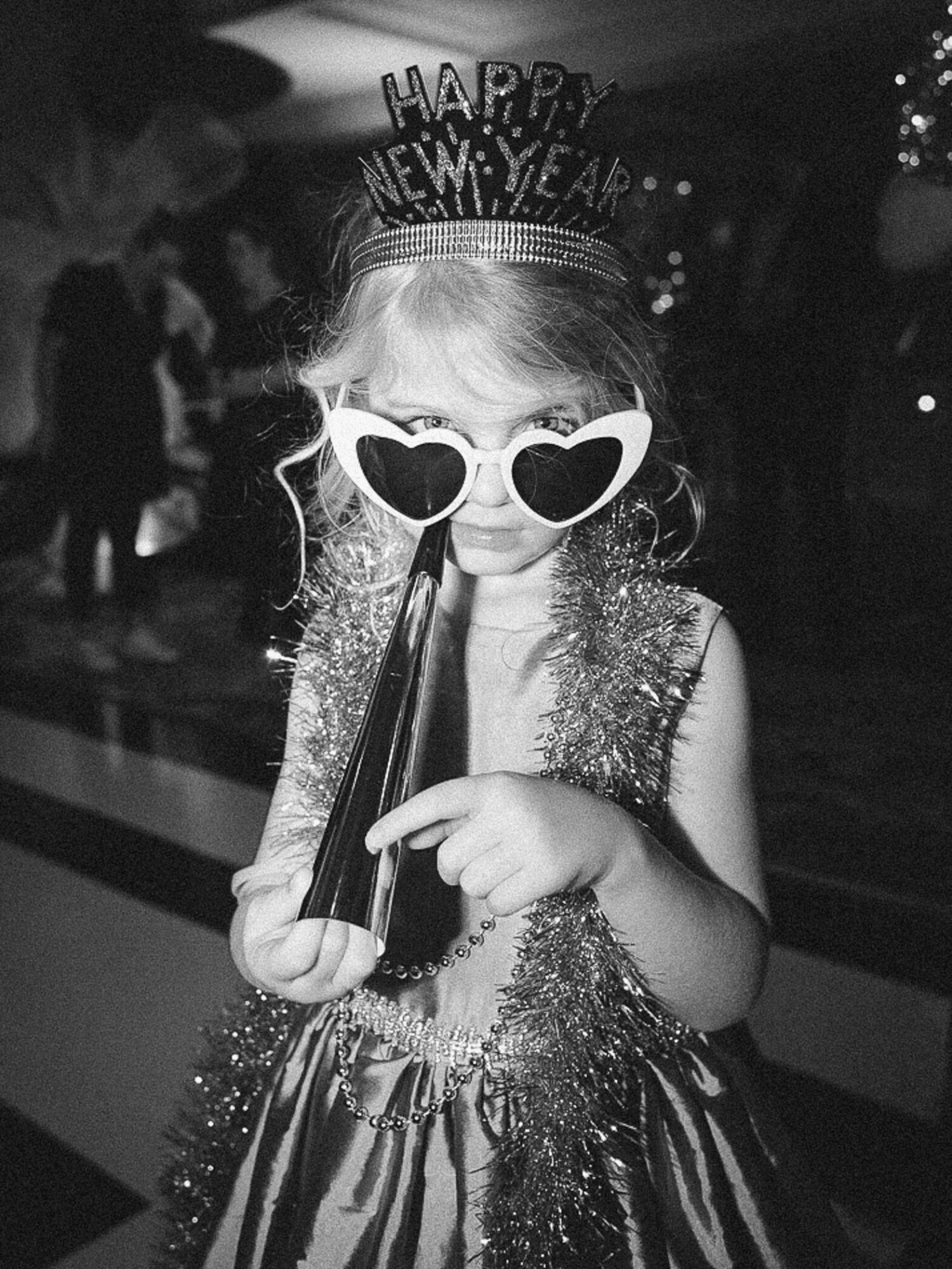 Child wearing heart-shaped sunglasses and a 'Happy New Year' headband, holding a party horn, in a festive setting.