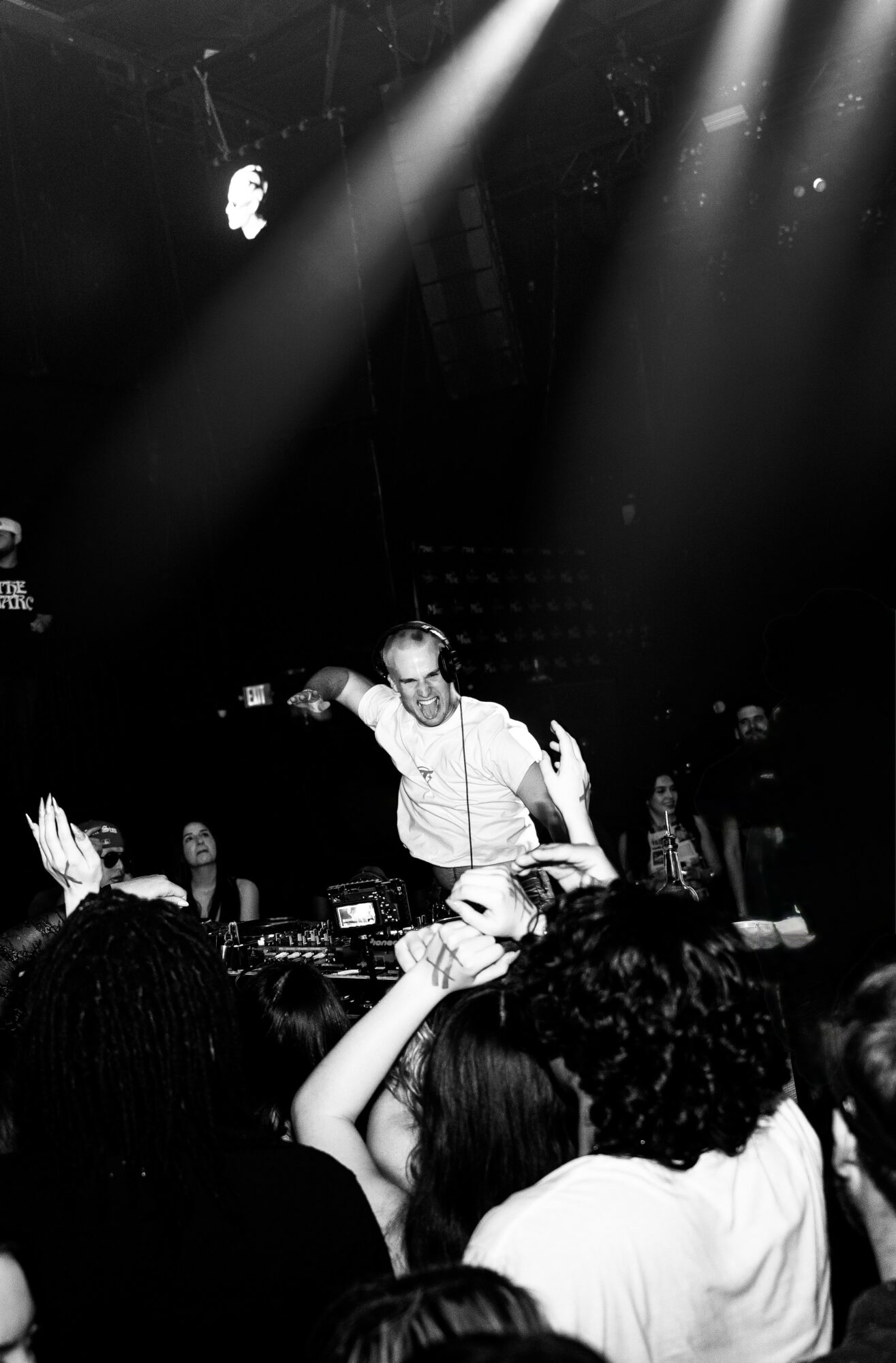 DJ performs on stage with audience in foreground, bright lights shining down, black and white image.