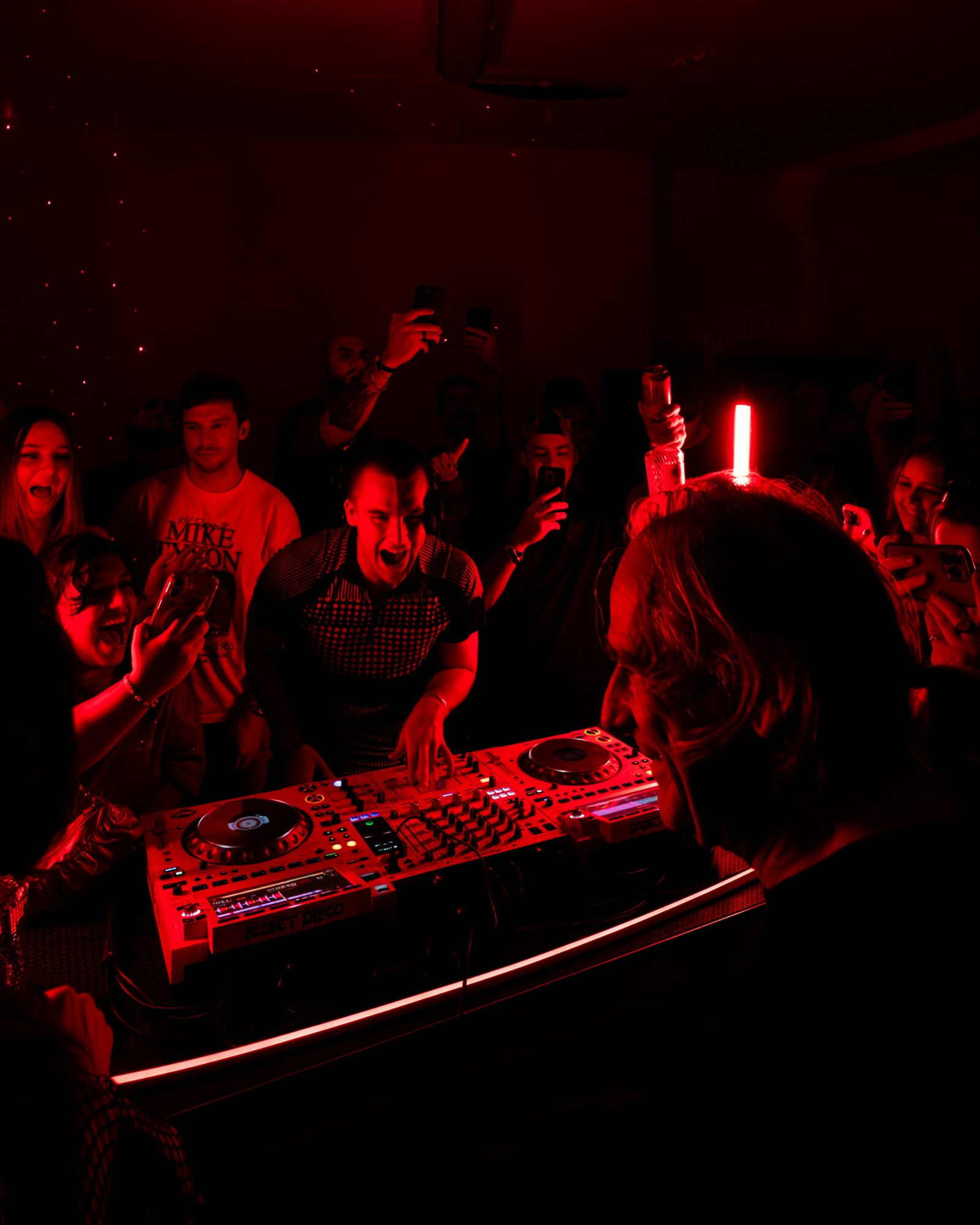 DJ performing at a dark party with red lighting, surrounded by people dancing and taking photos.