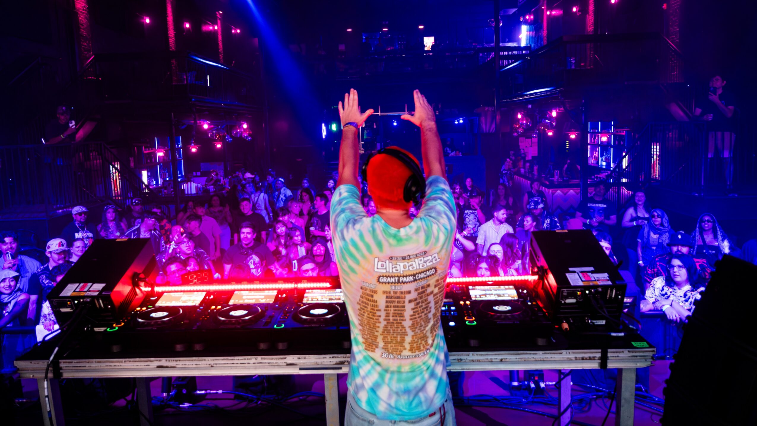 DJ with hands raised in a nightclub with colorful lights and a crowd in the background.