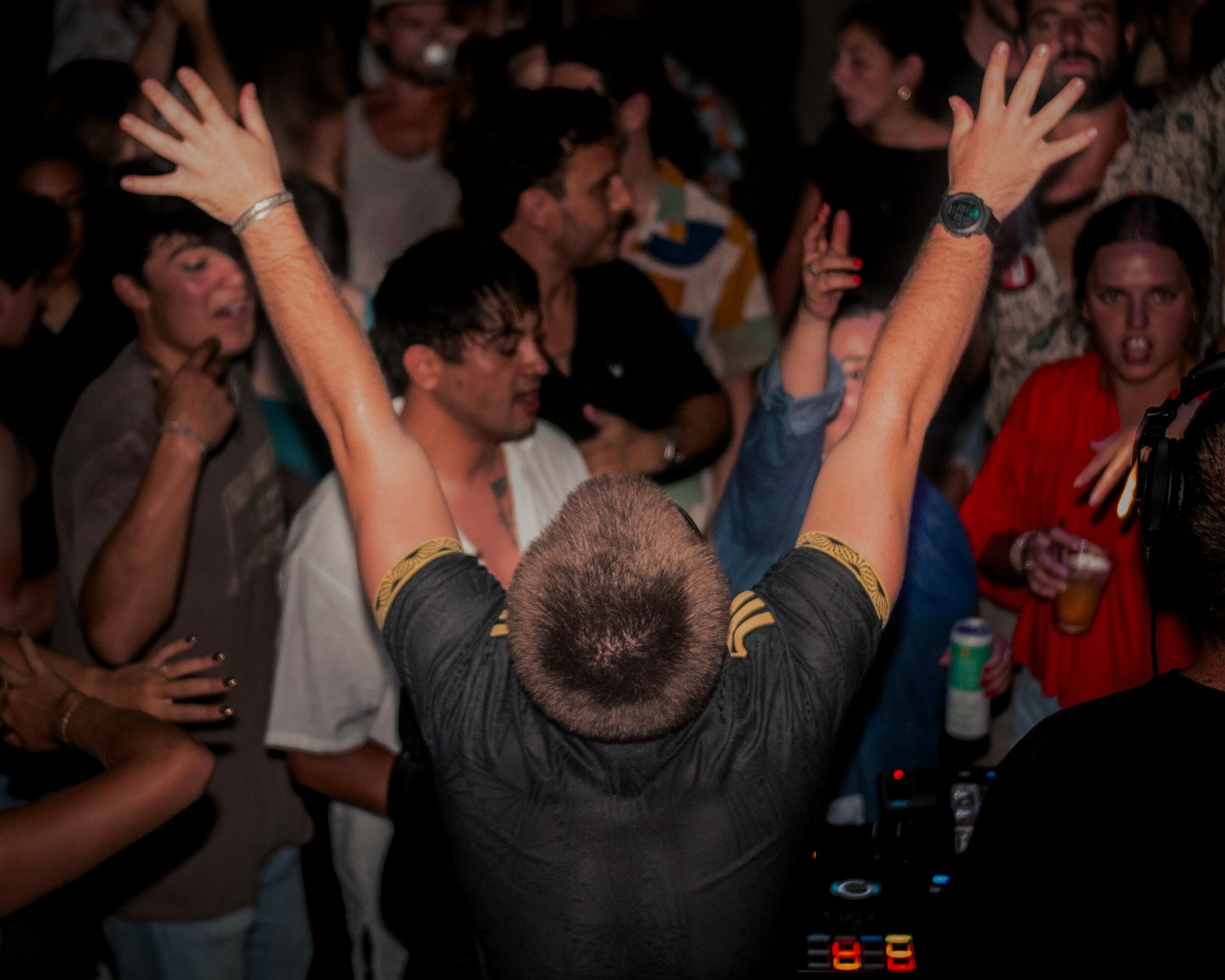 Person with arms raised facing a crowd at a party or event, with people dancing and socializing in the background.