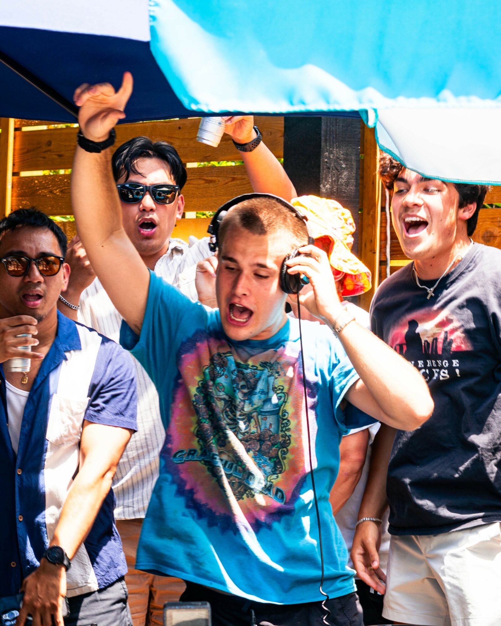 Group of young people outdoors, one wearing headphones, raising hand, others smiling and looking excited.