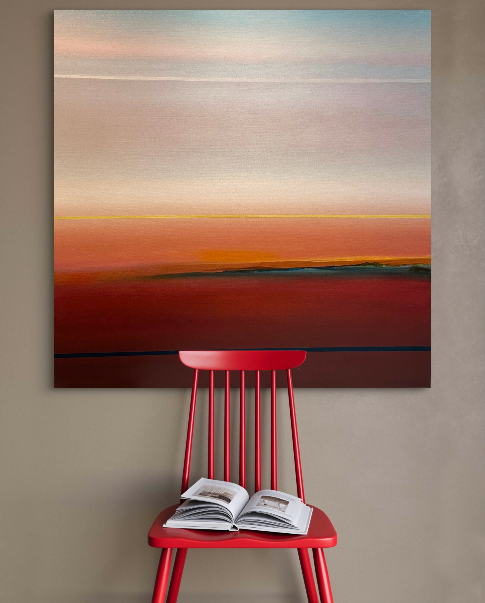 Red chair with open book in front of large abstract landscape painting, neutral wall background.