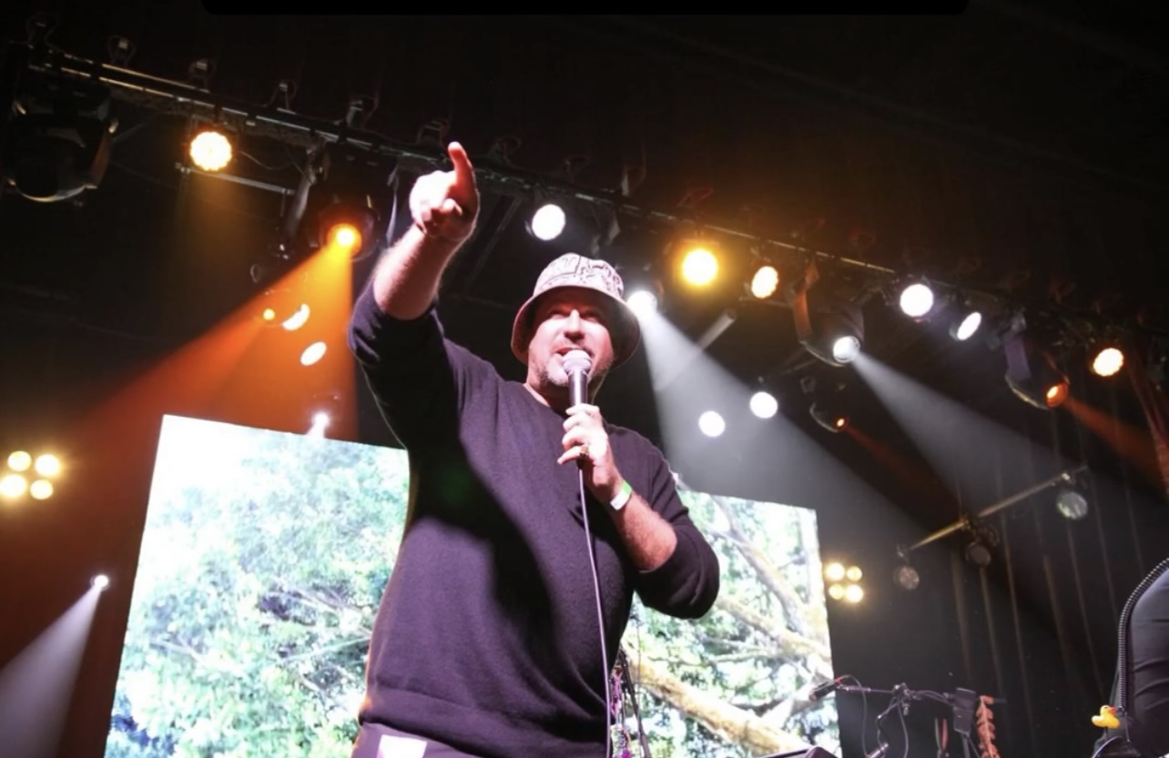 Person wearing a beanie and black sweater speaking into a microphone on stage with bright lights and screens behind.