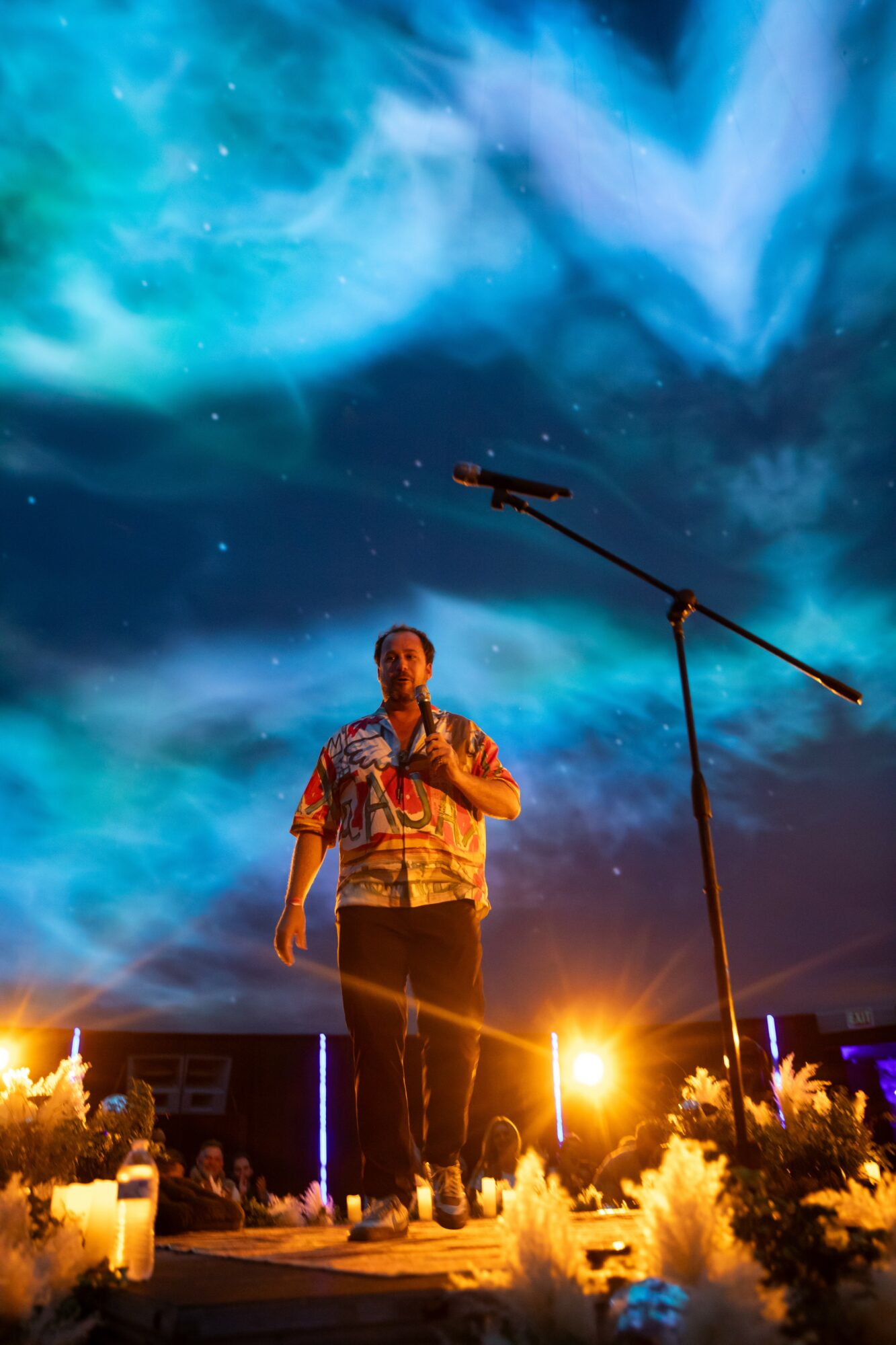 Person standing on stage under a colorful night sky with clouds and stars, microphone stand nearby, stage lights illuminate the scene.