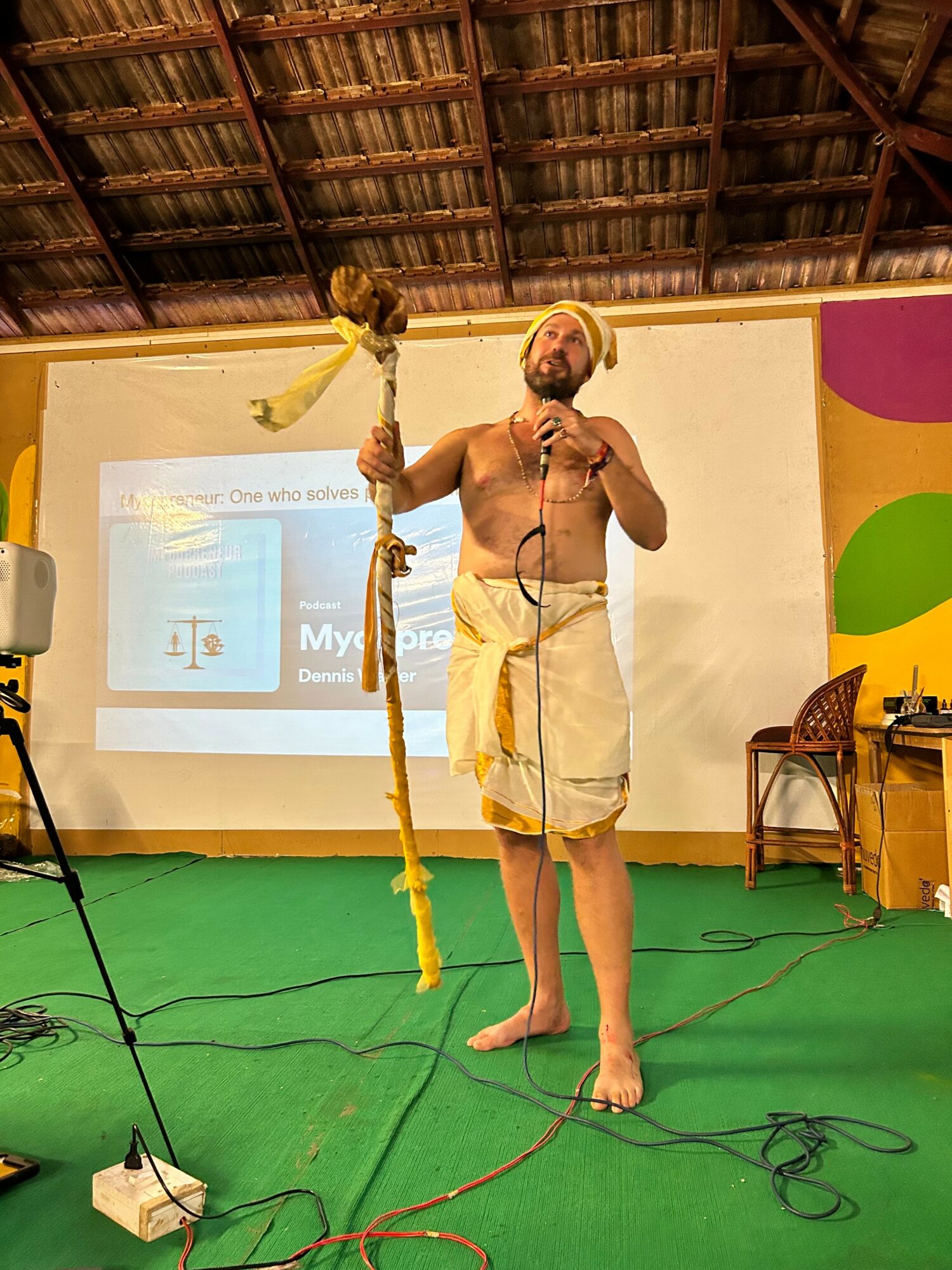 A shirtless man wearing a yellow turban and white dhoti stands on a green floor, holding a staff, in front of a screen and colorful wall.