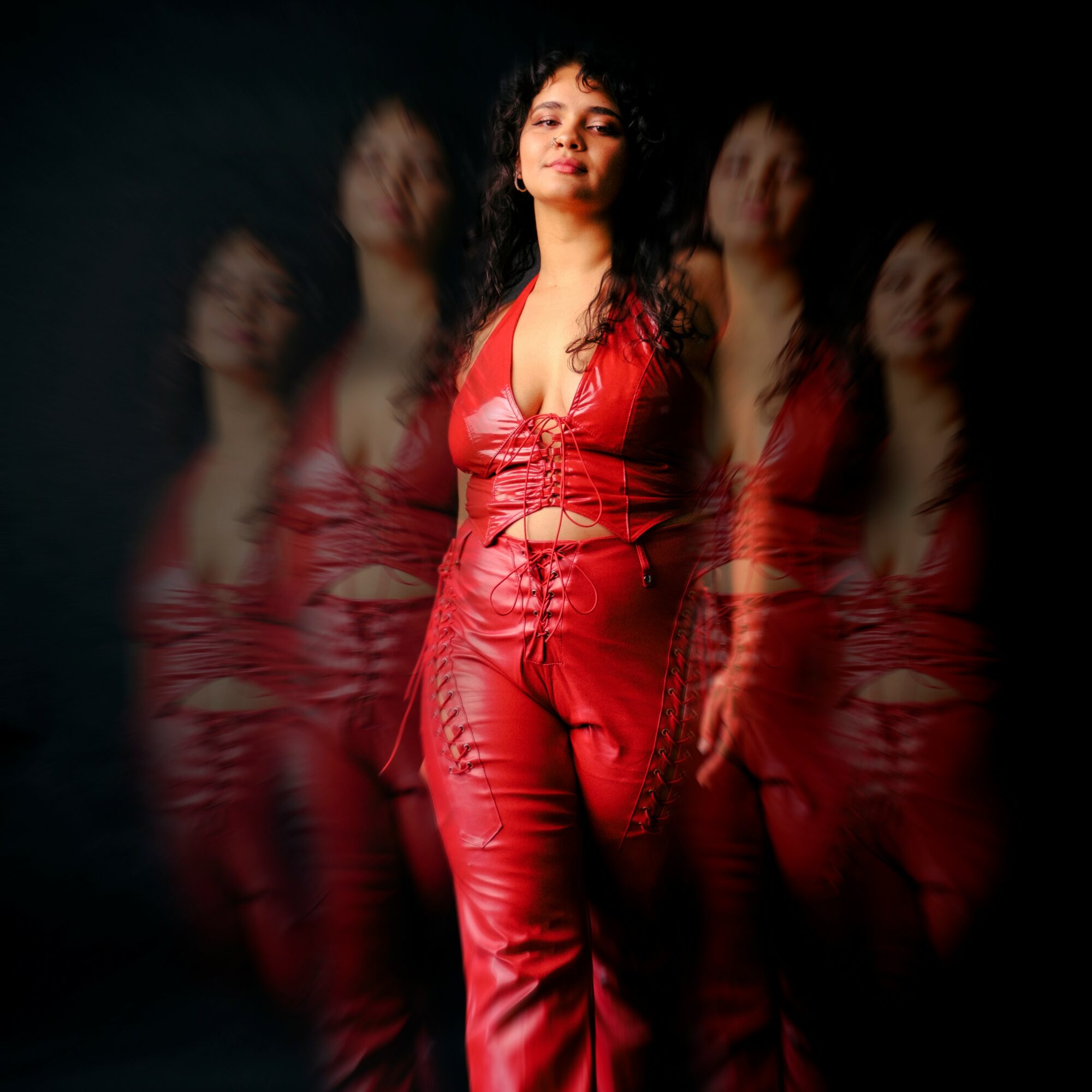Woman in red outfit standing with multiple blurred copies behind her, dark background.