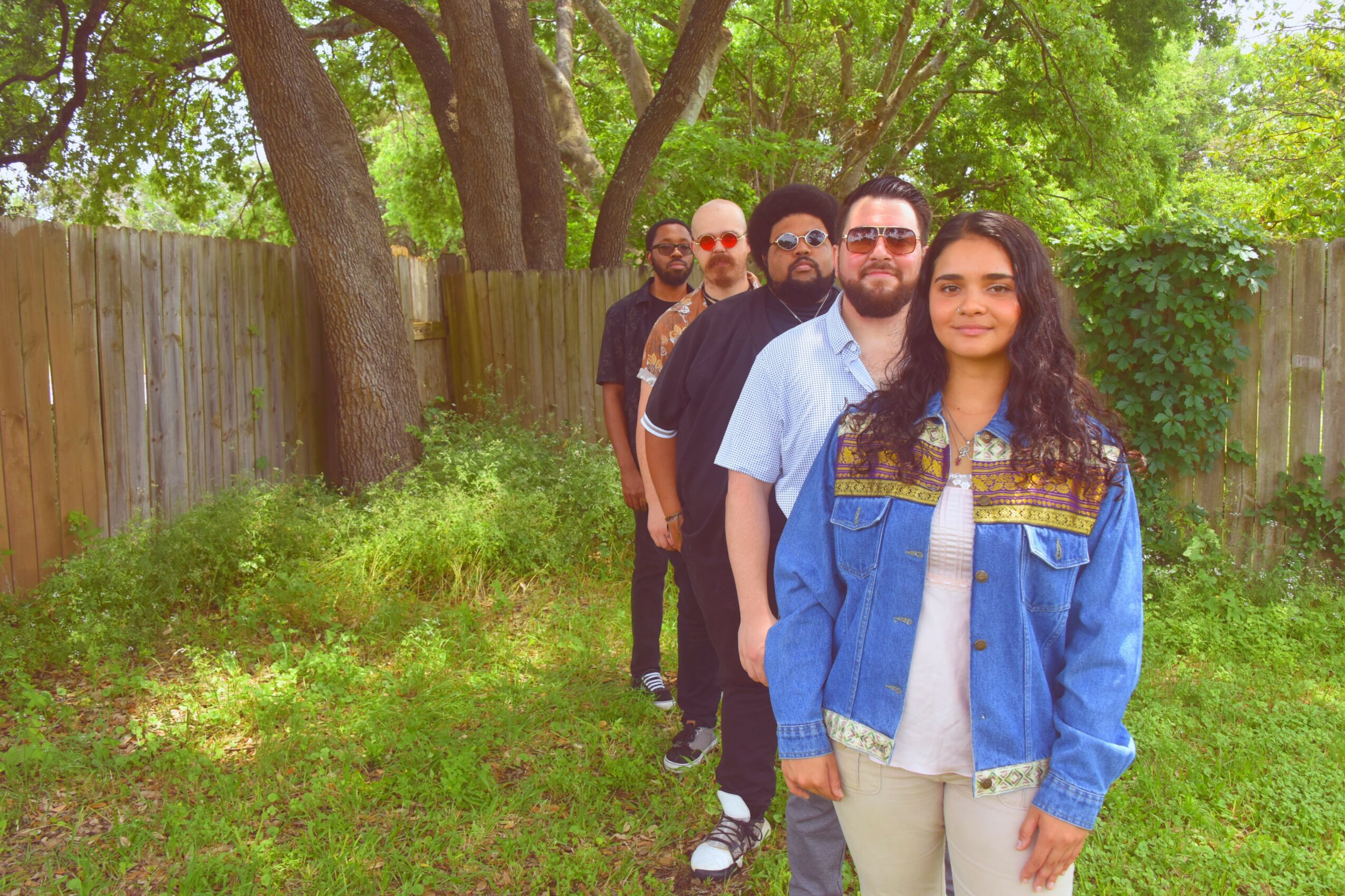 A group of six people standing in a line outdoors, with trees and a wooden fence in the background.