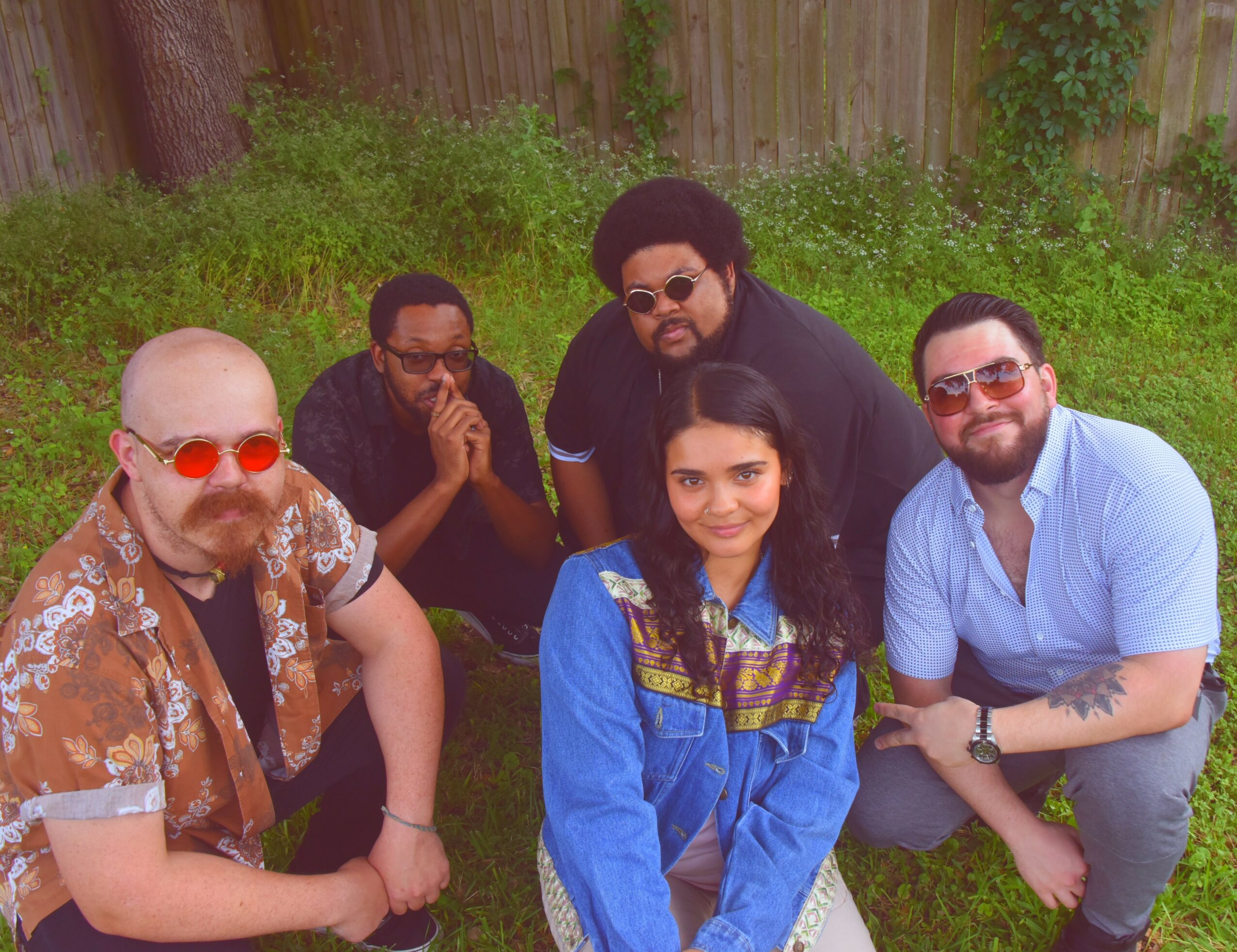 Five people outdoors, standing and crouching, with greenery and a wooden fence in the background.