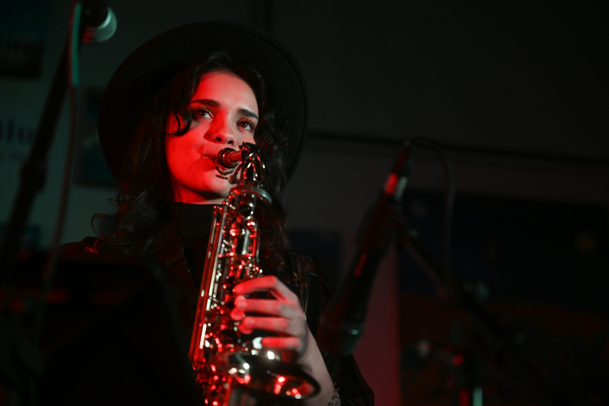 Woman with dark hair, wearing a hat, holding a saxophone, in a dimly lit setting.
