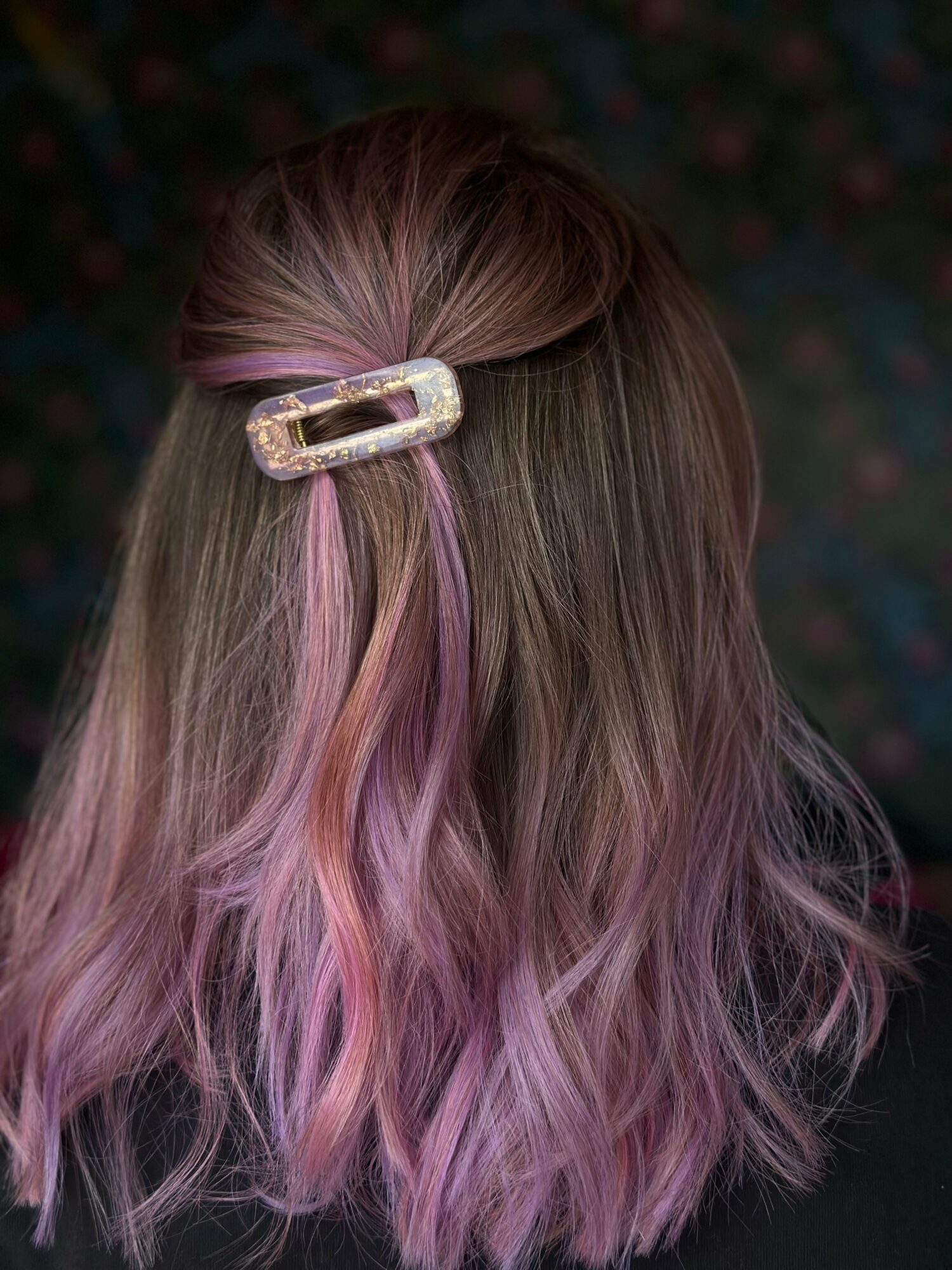 Back of a person's head with long, wavy hair dyed pink and purple, held with a decorative hair clip.