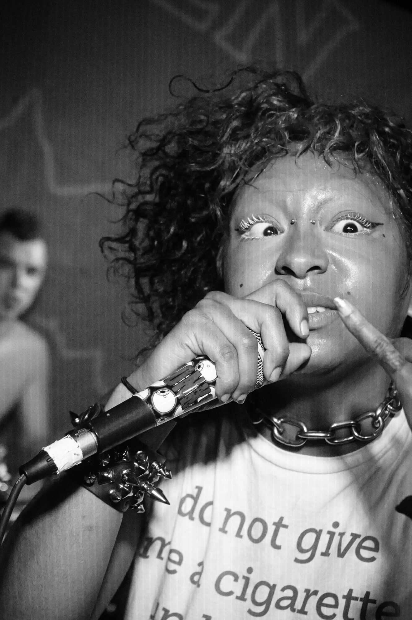 Person with curly hair holding a microphone close to their mouth, wearing a chain necklace and a T-shirt with text.