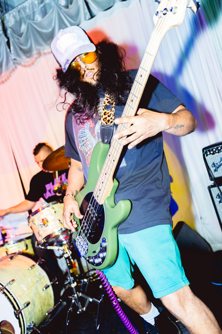 Person playing green electric bass guitar, wearing sunglasses, a cap, and casual clothing, with a drum set in the background.