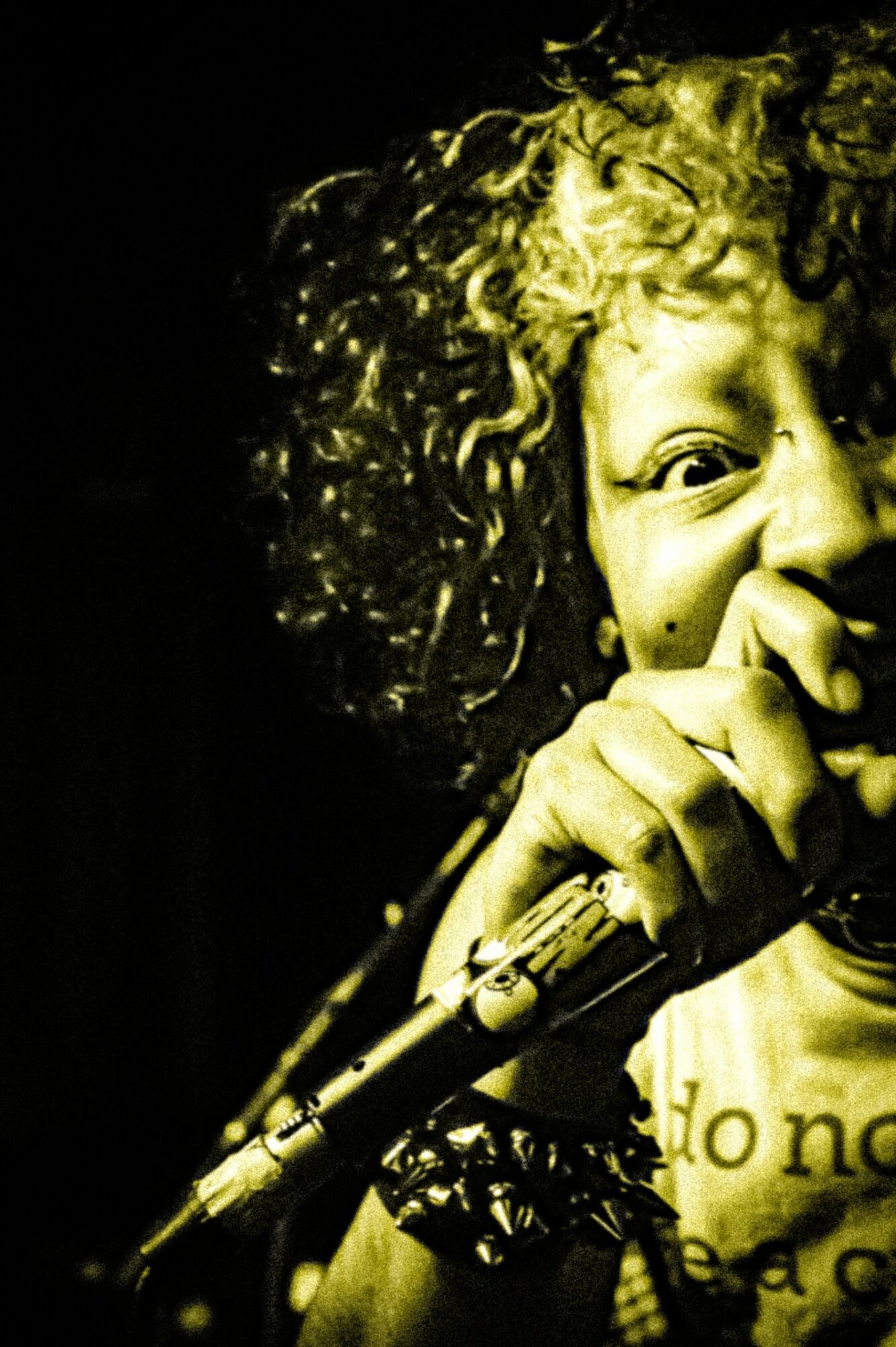 Close-up of a person with curly hair holding a microphone, smiling, with a dark background.