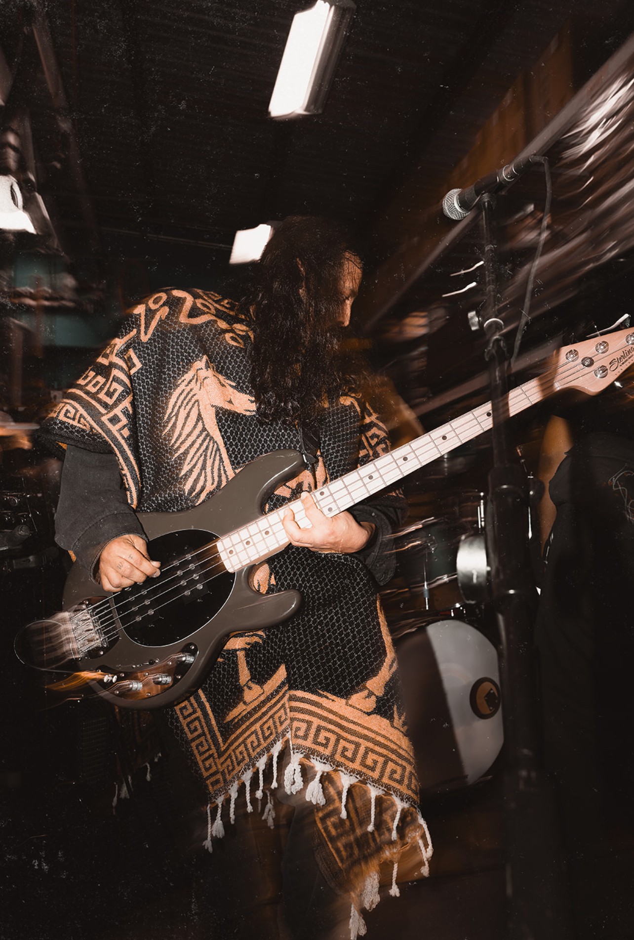 Person playing guitar on stage with dark background and stage lighting, wearing patterned clothing, holding a black guitar.