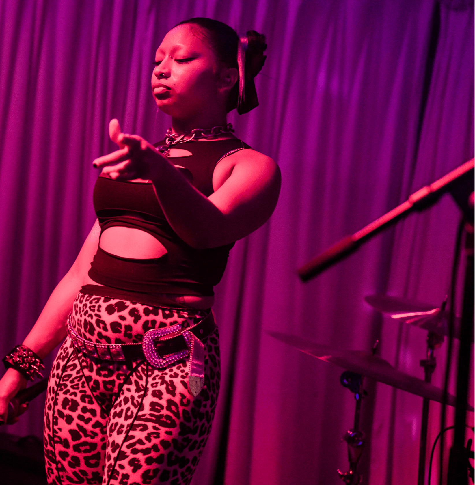 A woman performs on stage with purple curtains in the background, wearing a black top and leopard print pants.