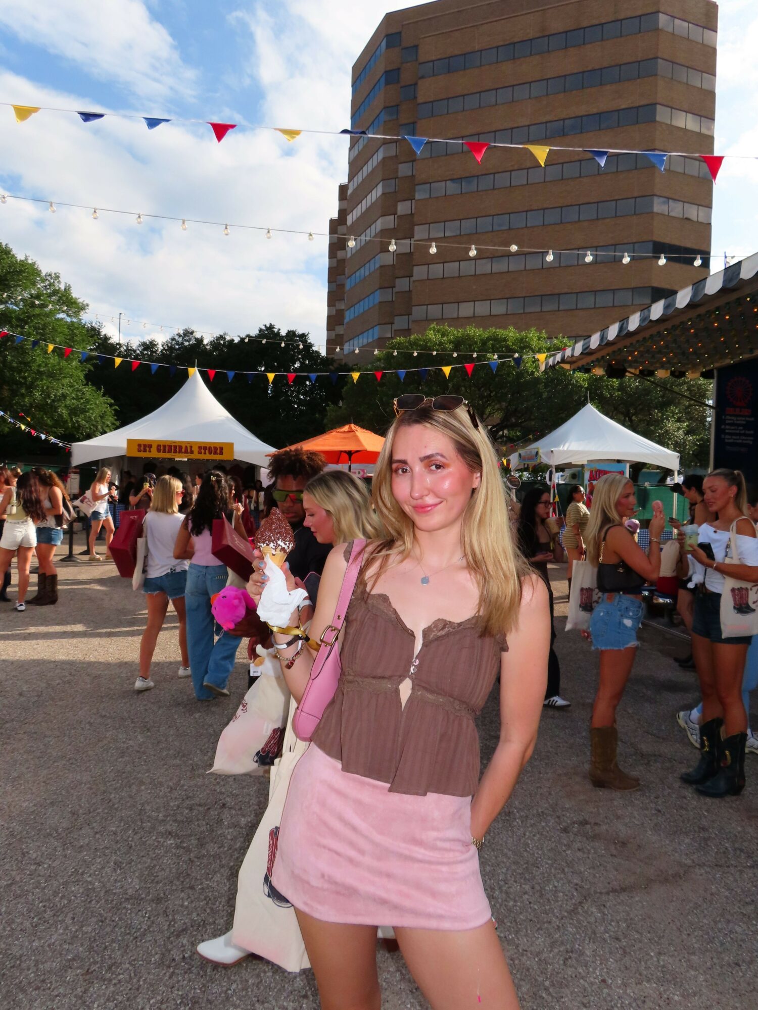 Young woman with blonde hair in a brown and pink outfit standing outdoors at a festival with tents and people in the background.