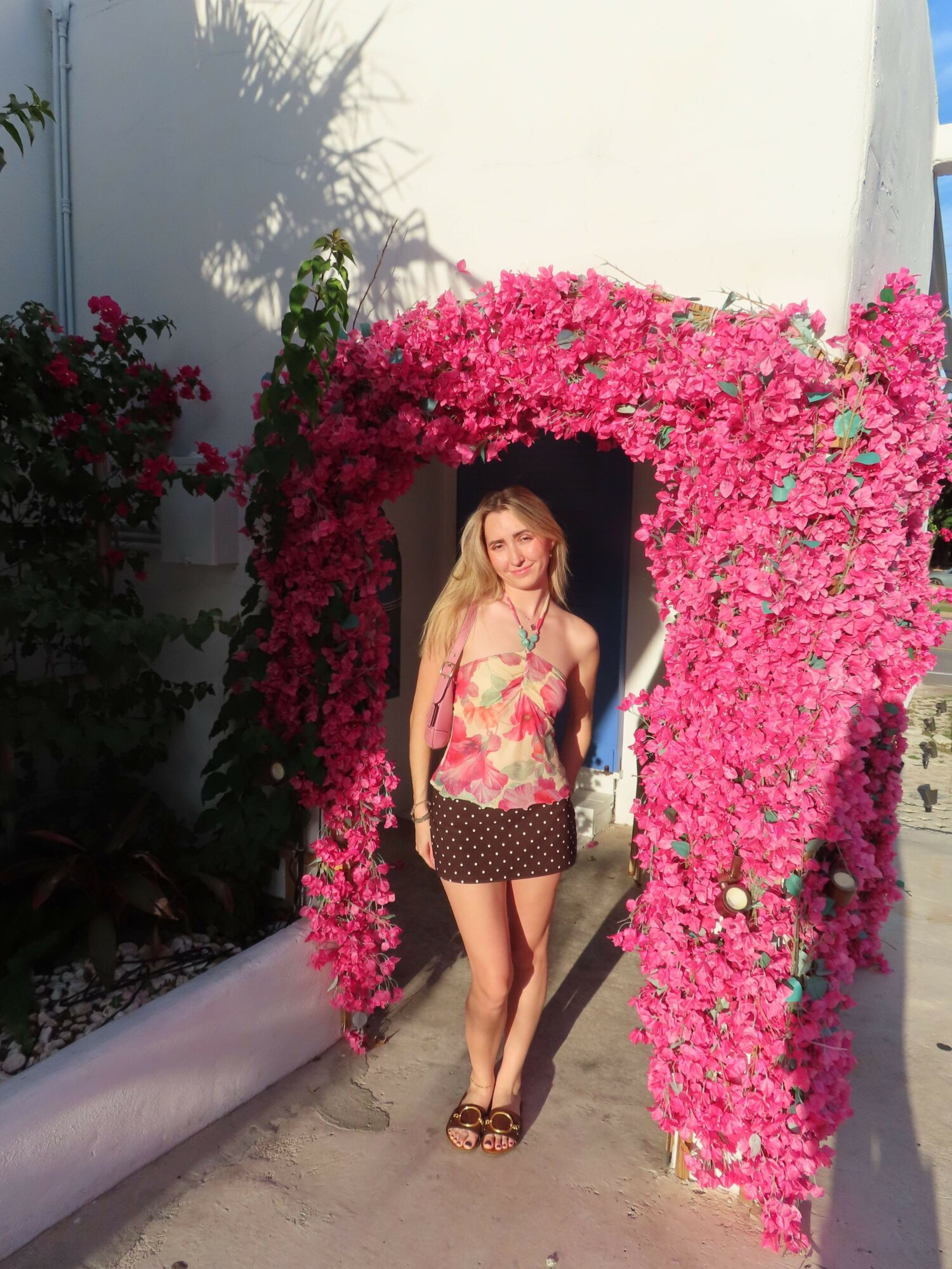Young woman standing in front of pink flowering arch, wearing floral top and dark shorts, smiling outdoors.