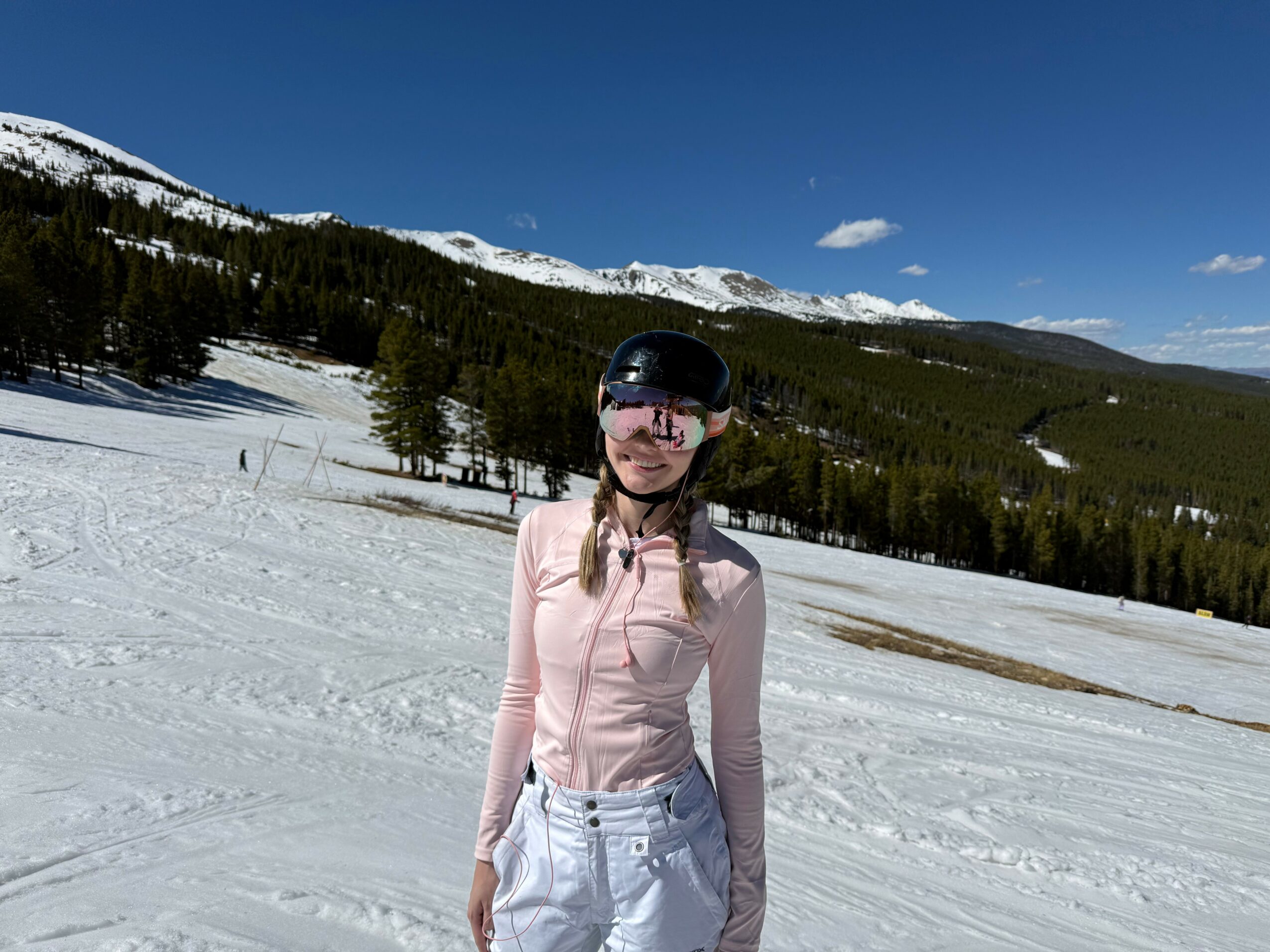 Girl wearing a helmet and sunglasses standing on snow with mountains and trees in the background.