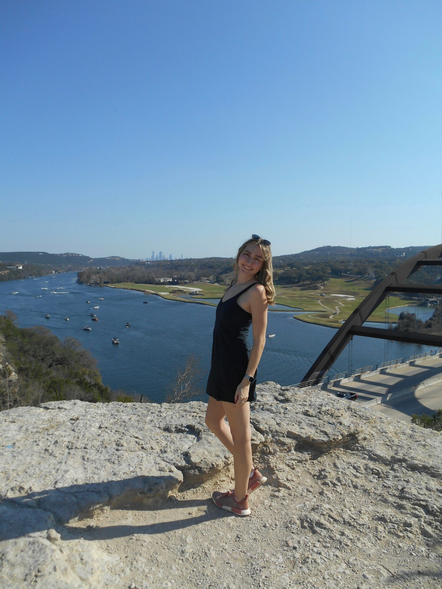 Young woman in black dress standing on rocky surface with water and landscape in background.
